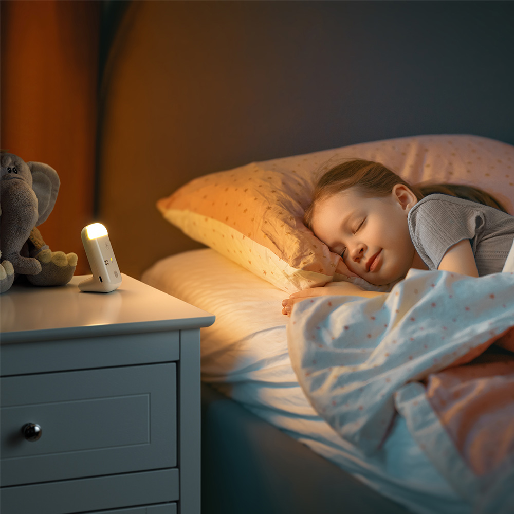 Un niño duerme plácidamente en la cama. Una mesita de noche con una lámpara y un peluche está al lado de la cama. La habitación está tenue.