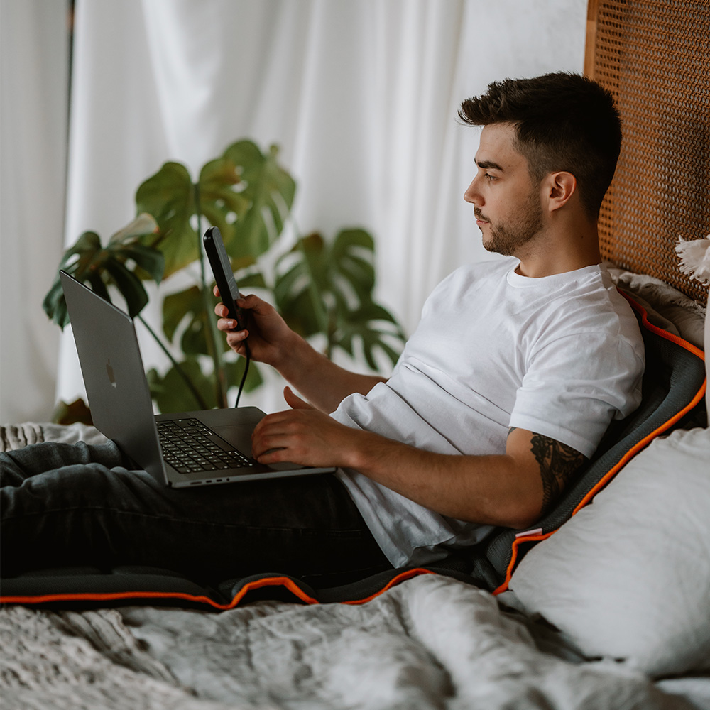 Ein Mann sitzt auf einem Bett und hält ein Telefon und einen Laptop. Hinter ihm ist eine Pflanze. Der Mann trägt ein weißes T-Shirt und dunkle Hosen.