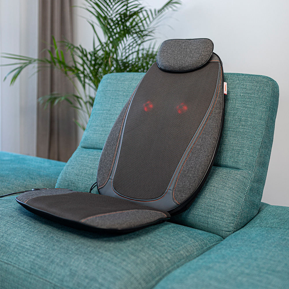 Ein Massagekissen auf einem türkisfarbenen Sofa. Es hat eine graue Netzoberfläche mit roten Lichtern. Im Hintergrund sind eine Pflanze und ein Vorhang.