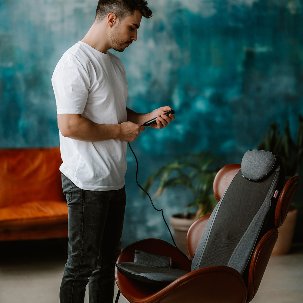 Ein Mann in einem weißen T-Shirt hält eine Fernbedienung und betrachtet einen Massagesessel. Der Stuhl ist braun und grau.