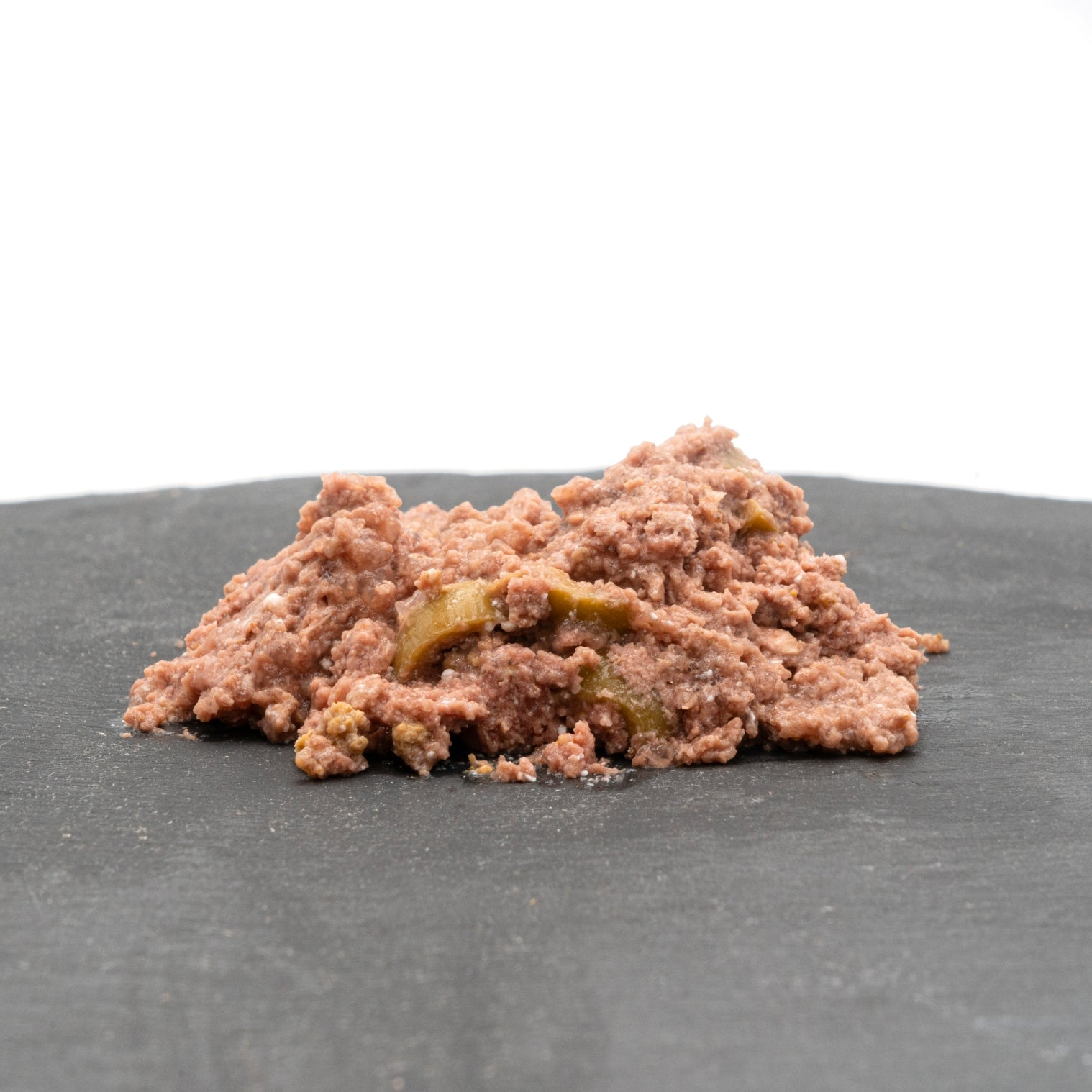 Un montón de comida húmeda para gatos sobre una superficie gris oscuro. La comida es de color marrón rosado con trozos visibles, algunos verdes. El fondo es blanco.