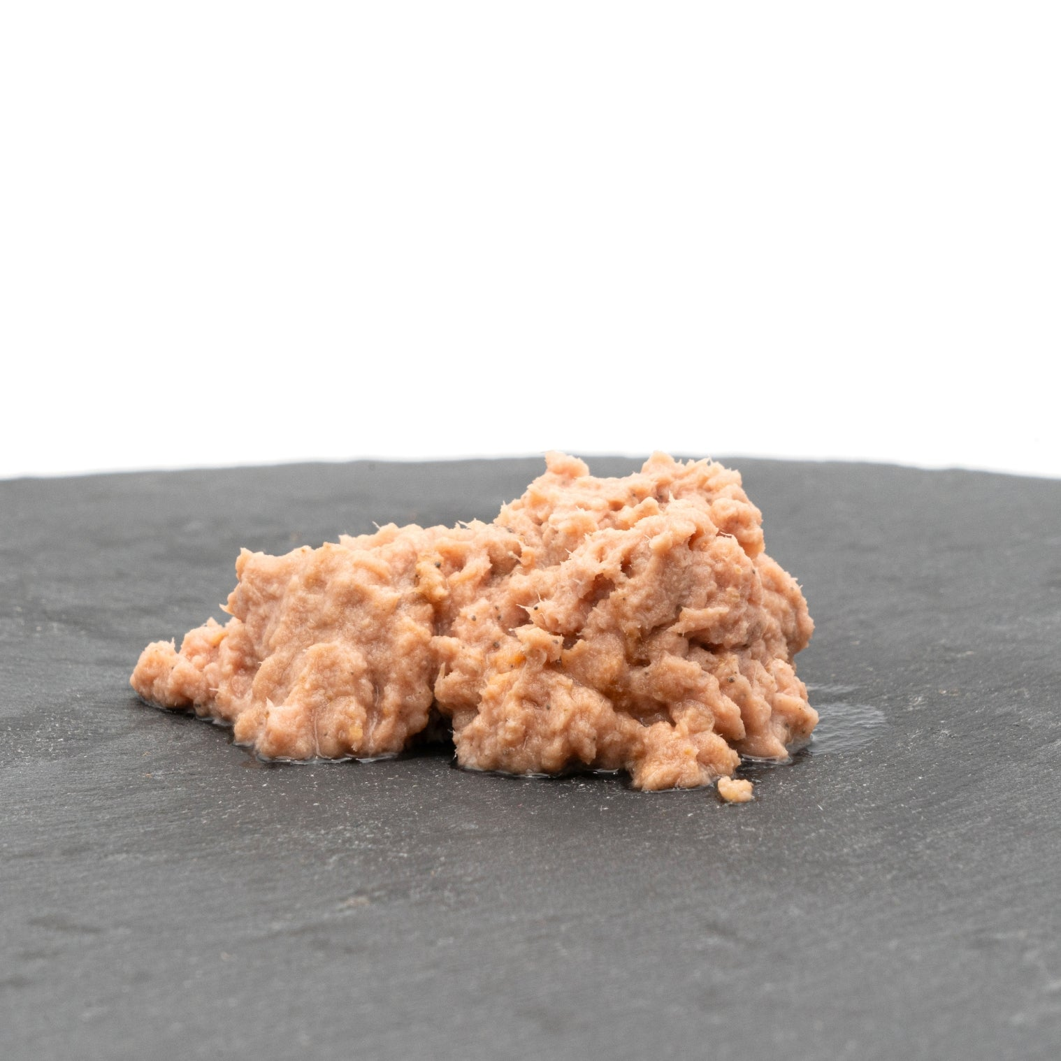 Un montón de comida desmenuzada de color rosa claro, posiblemente comida para gatos, descansa sobre una superficie gris oscura y texturizada, sobre un fondo blanco.