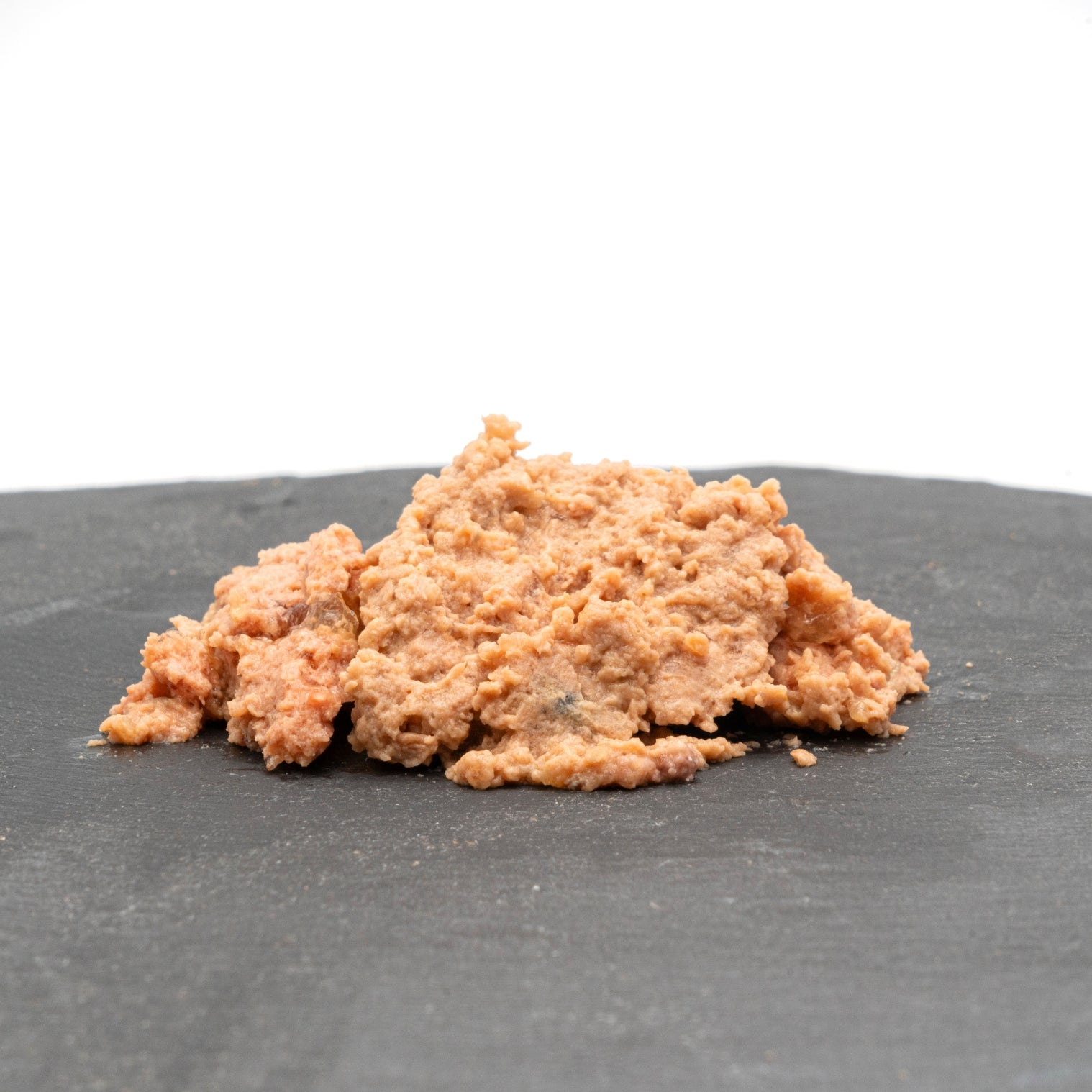 Un montón de comida de color marrón claro y texturizada está sobre una superficie gris oscuro, sobre un fondo blanco. La comida parece ser comida para gatos.