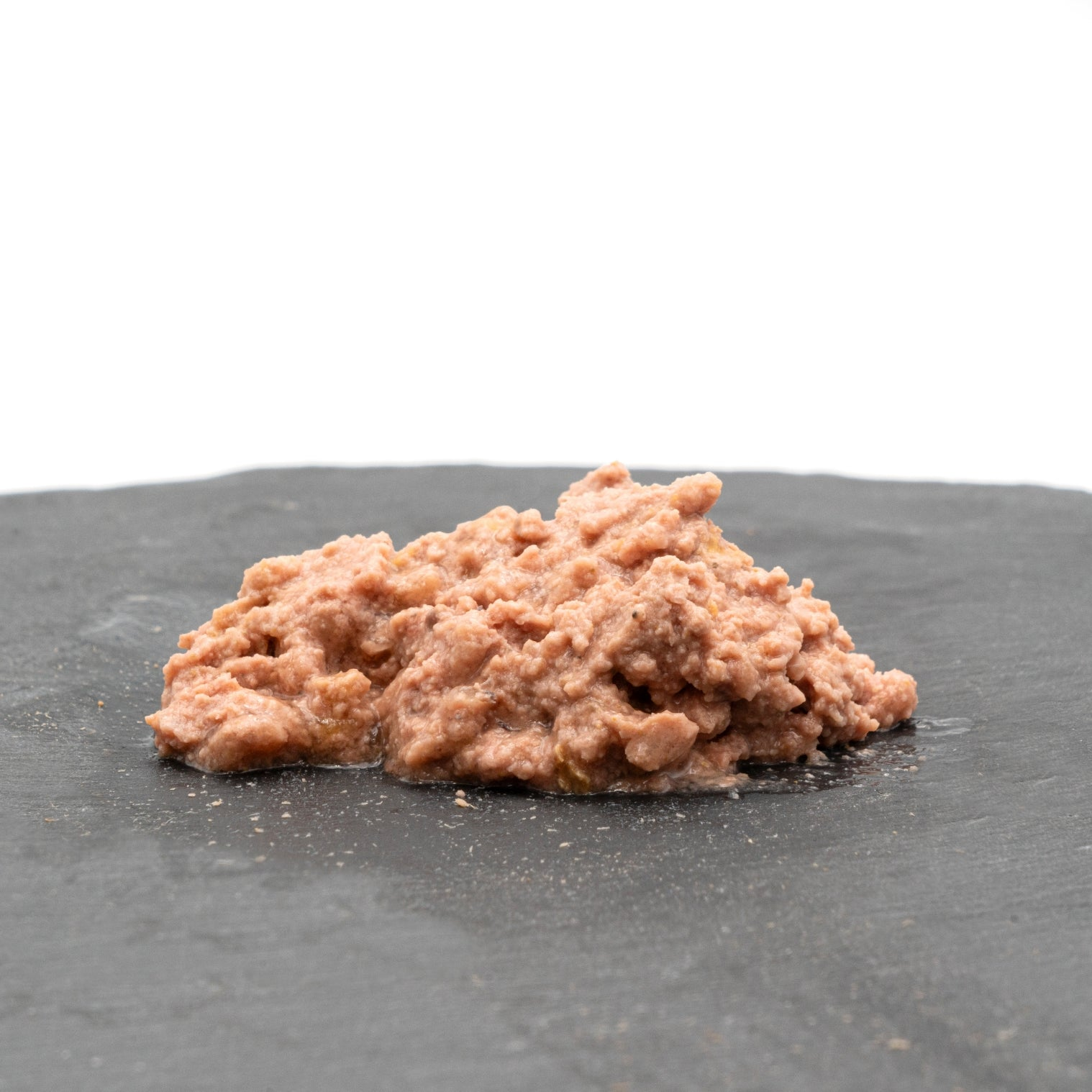 Un pequeño montón de comida húmeda para mascotas sobre una superficie gris oscuro. La comida es de color marrón rosado, con un aspecto húmedo y ligeramente texturizado.