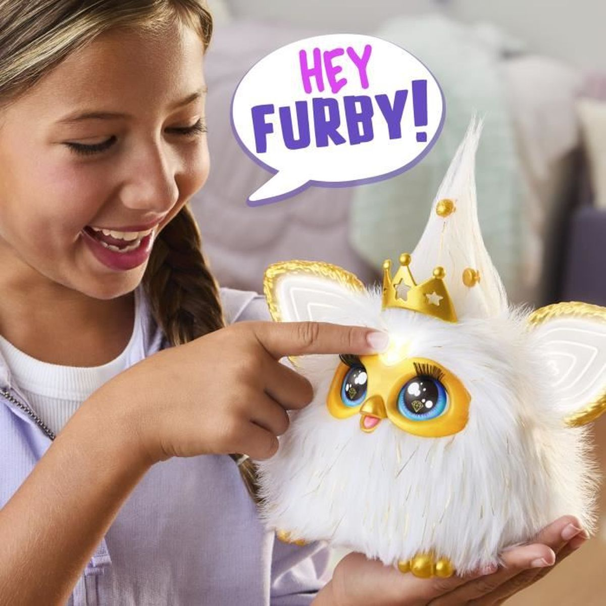 Ein Mädchen lächelt und zeigt auf ein weißes Furby-Spielzeug mit goldener Krone. Sprechblase sagt "HEY FURBY!". Furby hat goldene Akzente.
