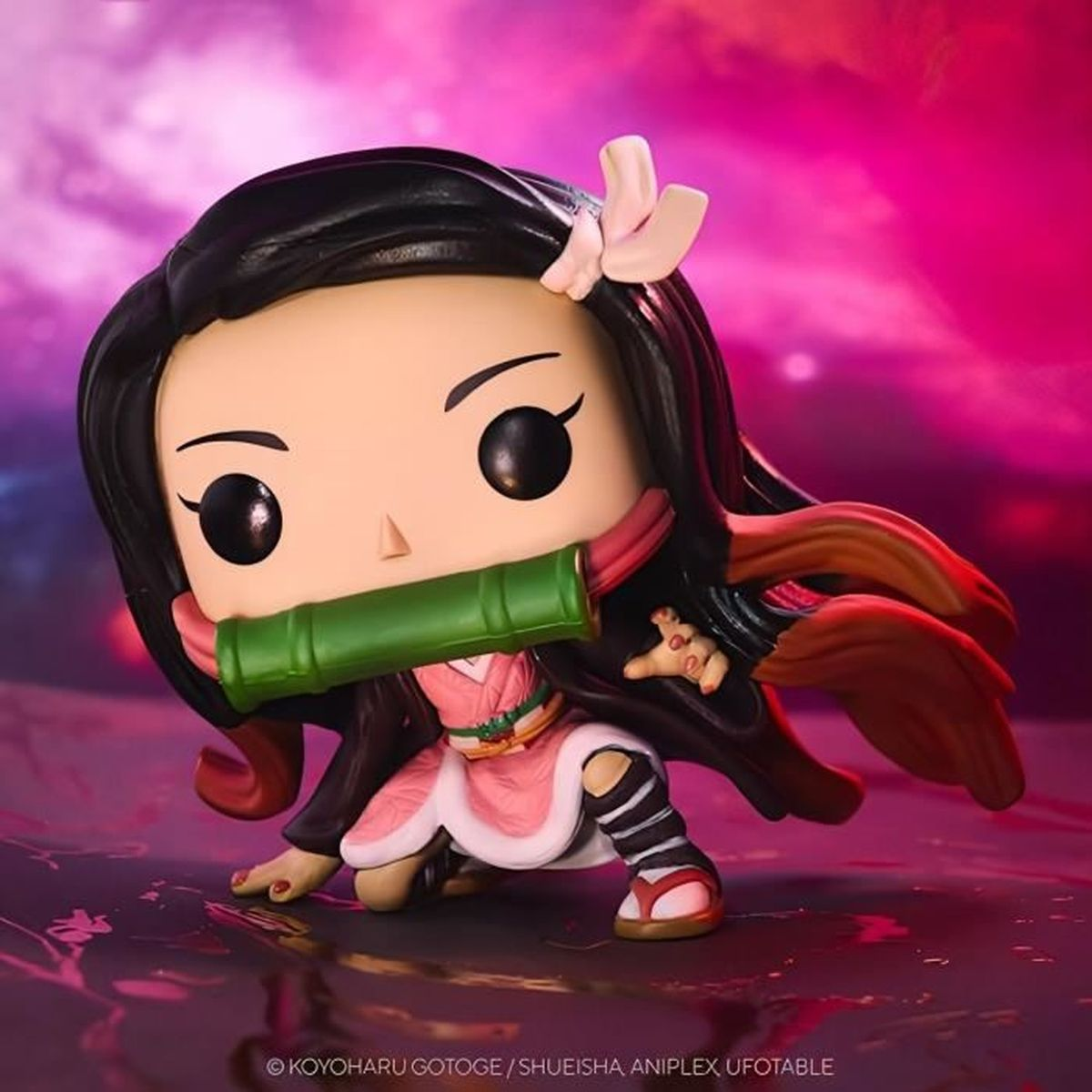Eine Funko Pop-Figur von Nezuko Kamado aus Dämonentöter, kniend. Sie hat einen Bambus-Mund, einen rosa Kimono und schwarze Haare. Der Hintergrund ist pink.