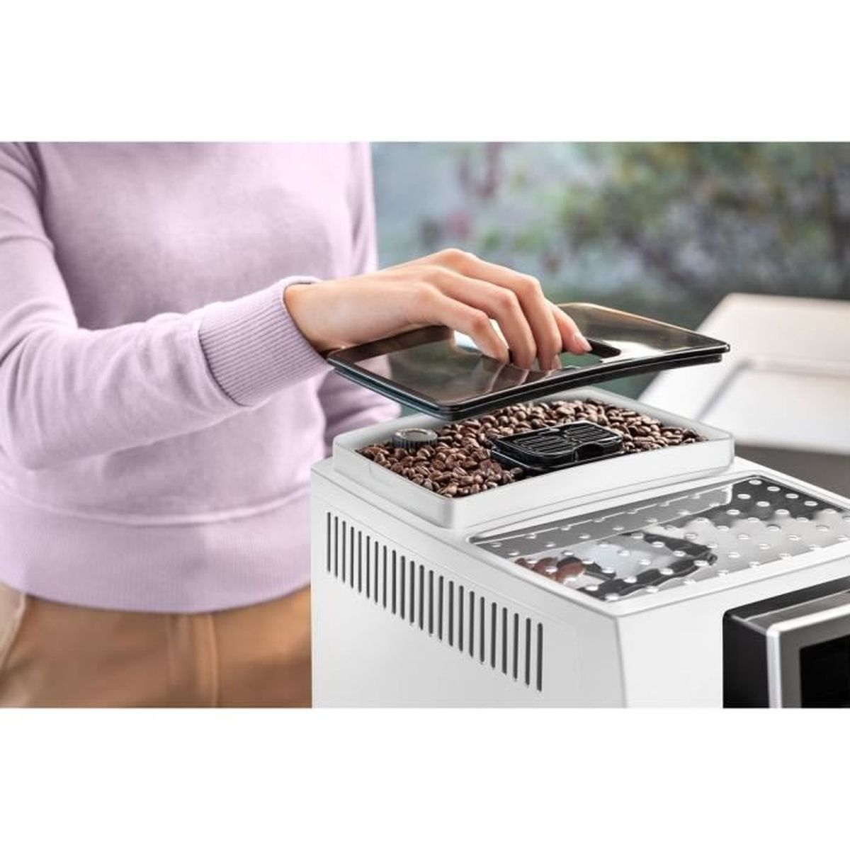 Una persona apre il coperchio del contenitore dei chicchi di una macchina da caffè. Il contenitore è pieno di chicchi di caffè. La macchina è bianca.