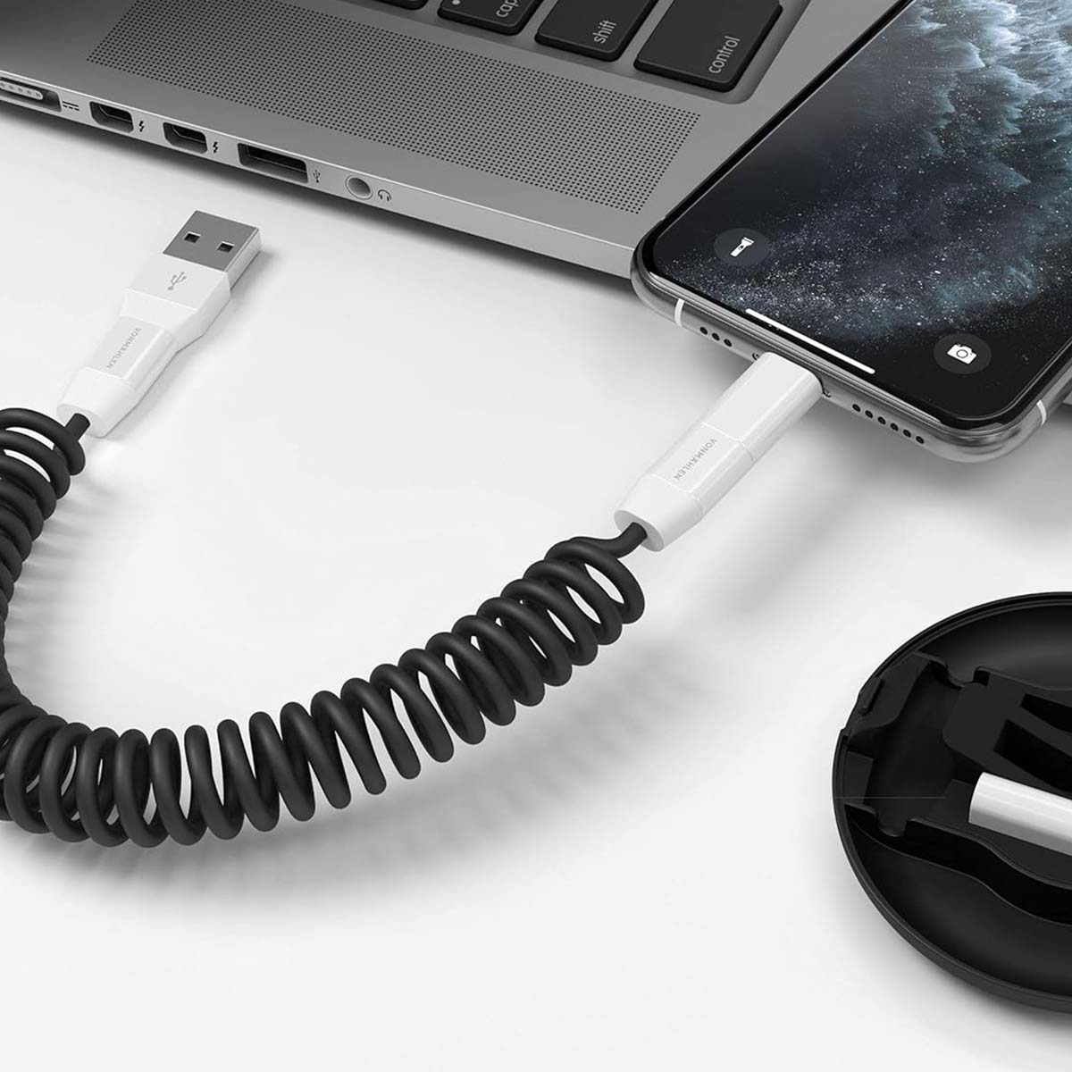 Un telefono è in carica con un cavo a spirale nero collegato a un laptop. Una penna bianca è vicino a una custodia nera.