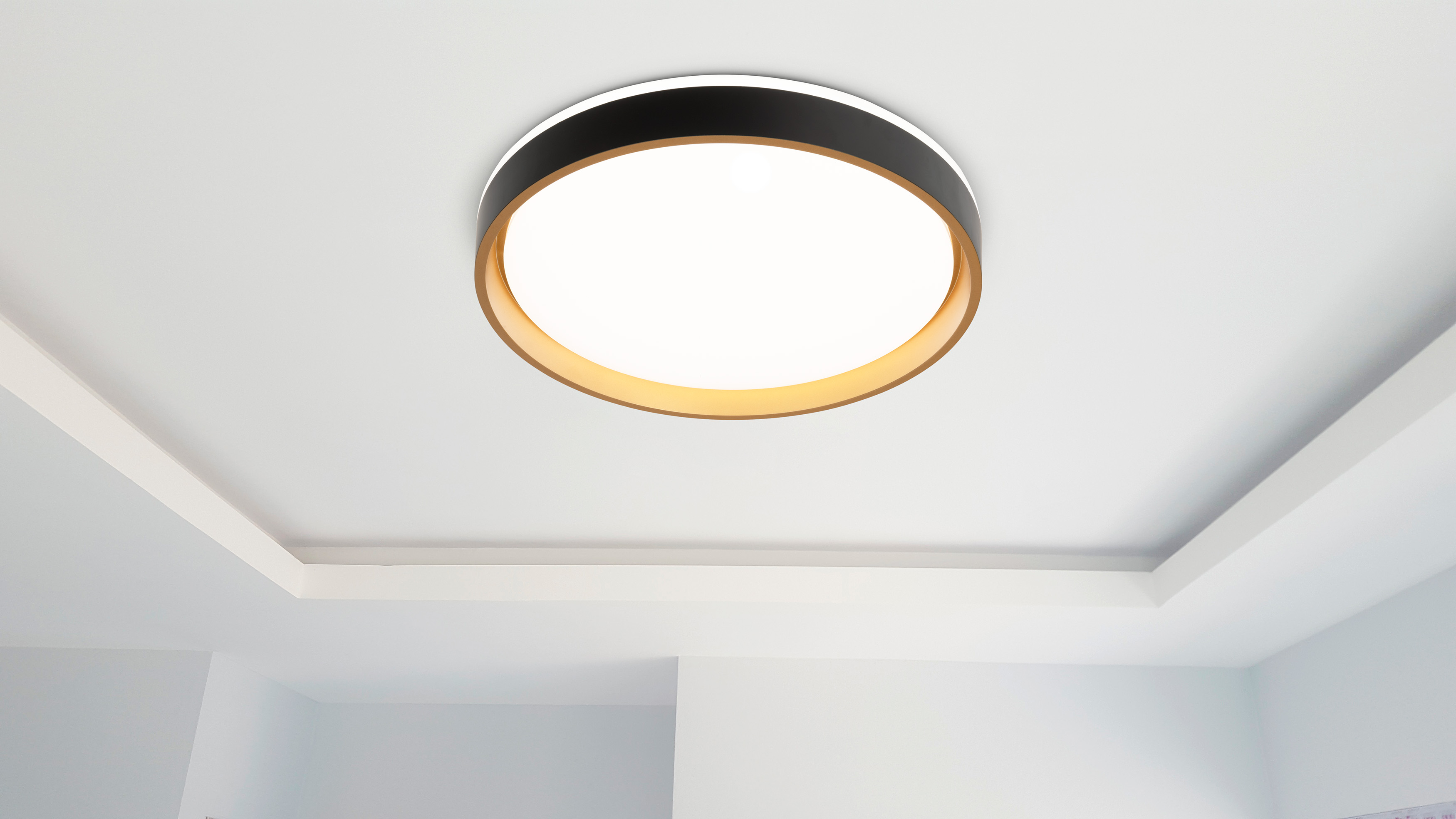Eine Deckenleuchte mit schwarzem Außenring und goldenem Innenring, die helles Licht ausstrahlt. Die Decke ist weiß.