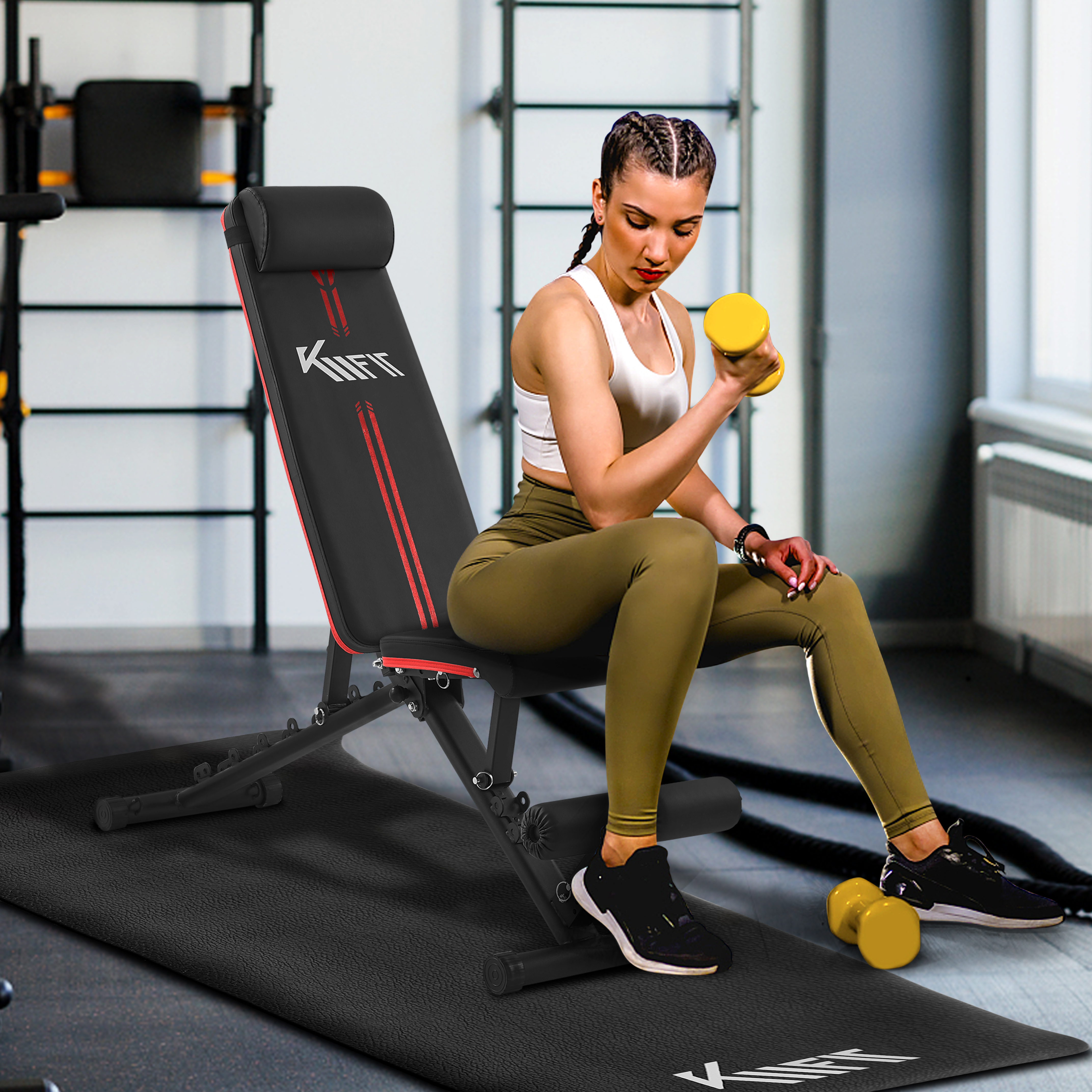 Eine Frau im Fitnessstudio, die eine gelbe Hantel hebt, während sie auf einer schwarz-roten Bank sitzt, mit weiteren Hanteln und einer Matte.