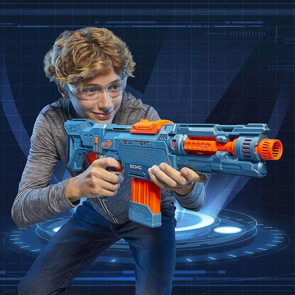 Ein Junge mit Brille hält eine blau-orangefarbene Spielzeugpistole und lächelt. Er trägt ein graues Hemd und Jeans. Der Hintergrund ist blau.