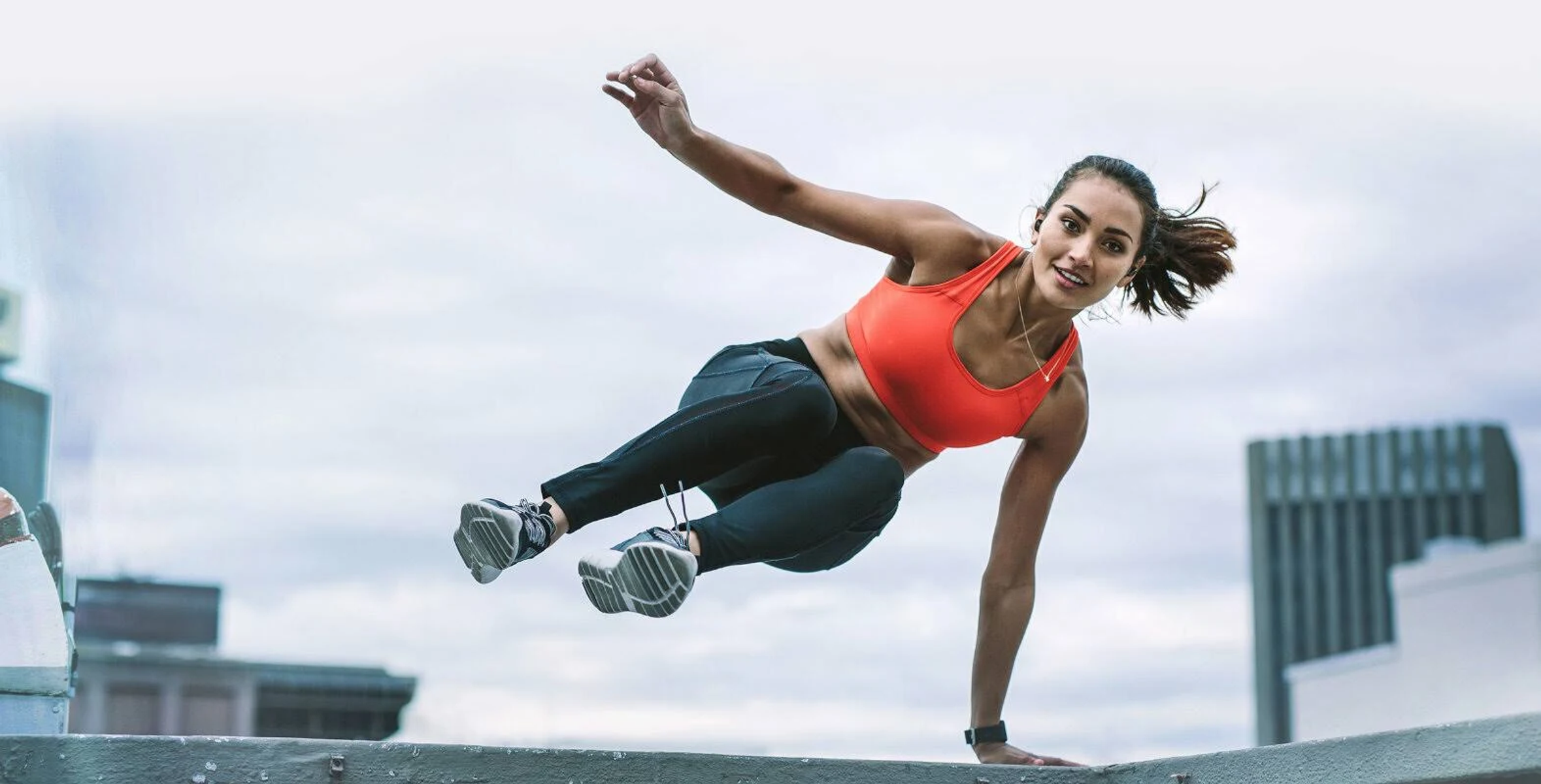Donna che fa parkour su un tetto. Indossa un reggiseno sportivo arancione e leggings neri. Edifici sullo sfondo.