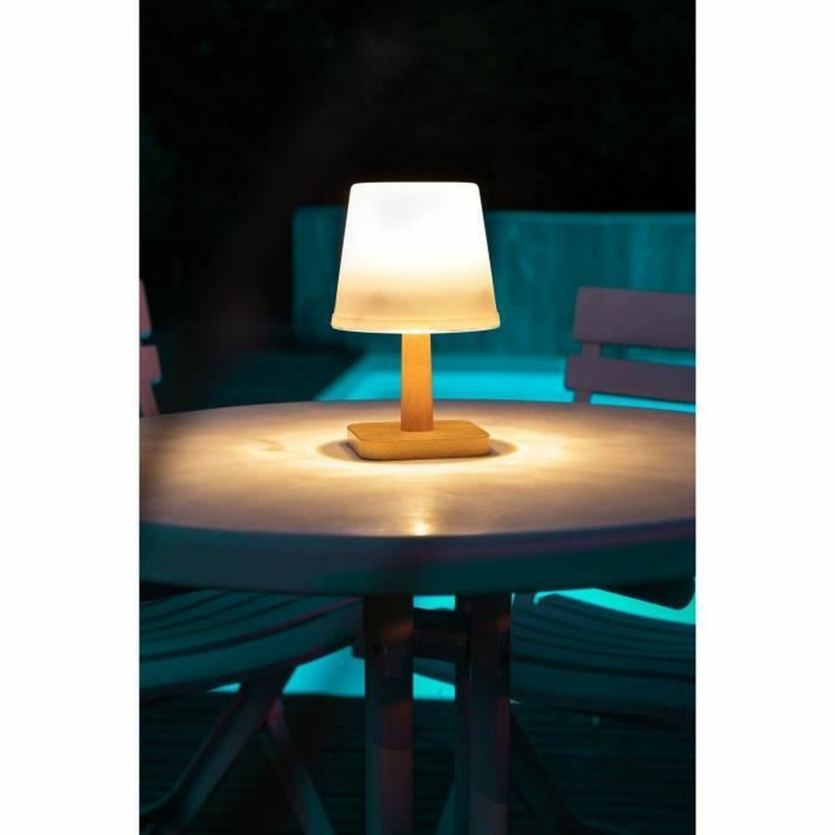 Eine beleuchtete Lampe mit weißem Lampenschirm steht auf einem Holztisch. Die Szene ist im Freien, mit Stühlen und dunklem Hintergrund.