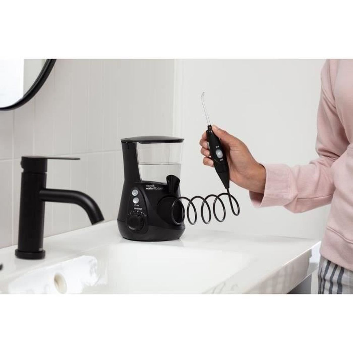 Ein schwarzes Munddusche auf einem weißen Waschbecken mit einem schwarzen Wasserhahn. Eine Person hält die Munddusche. Die Szene spielt in einem Badezimmer.
