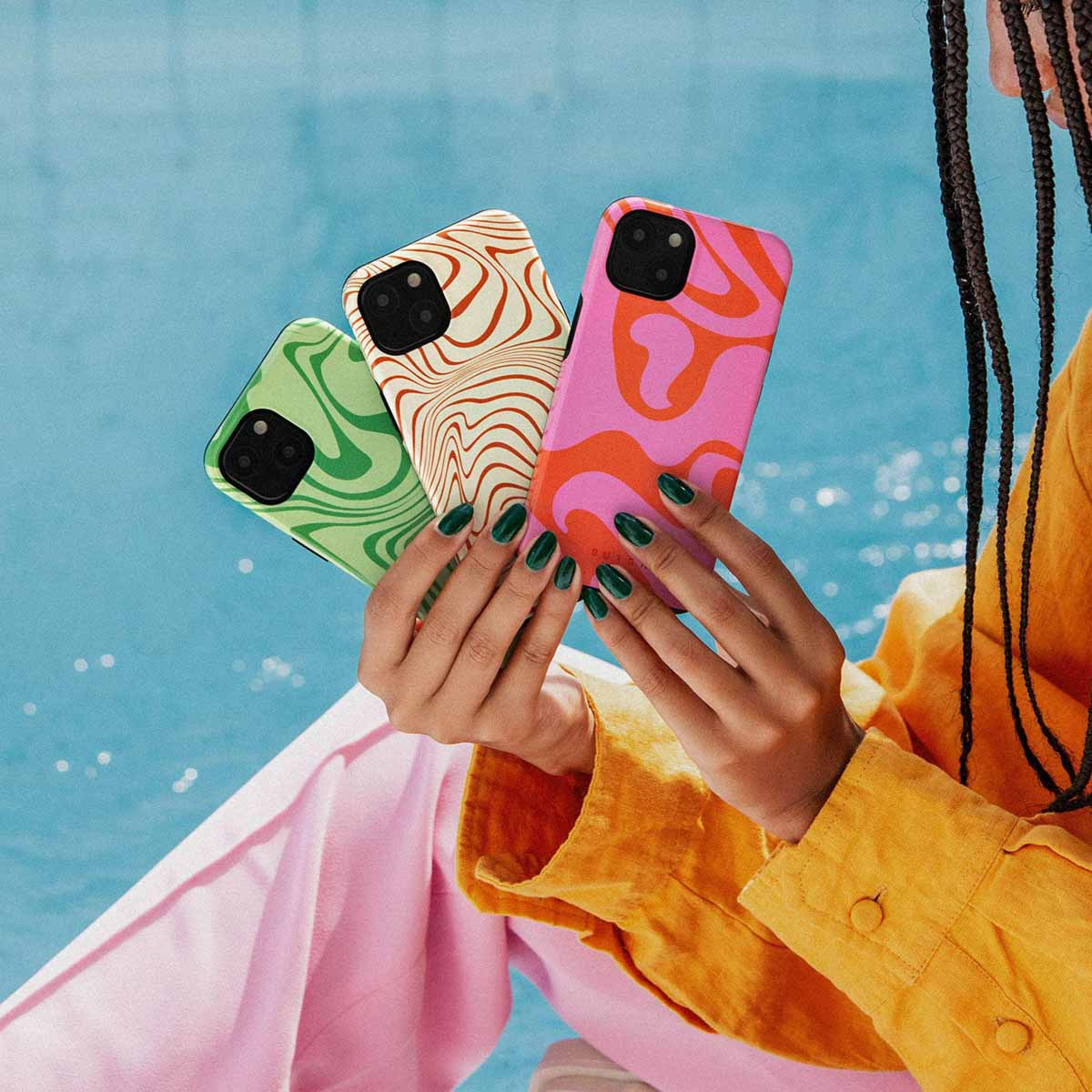 Een persoon houdt drie telefoonhoesjes met patronen vast. De hoesjes zijn groen, beige en roze. De persoon draagt een gele blouse en een roze broek.