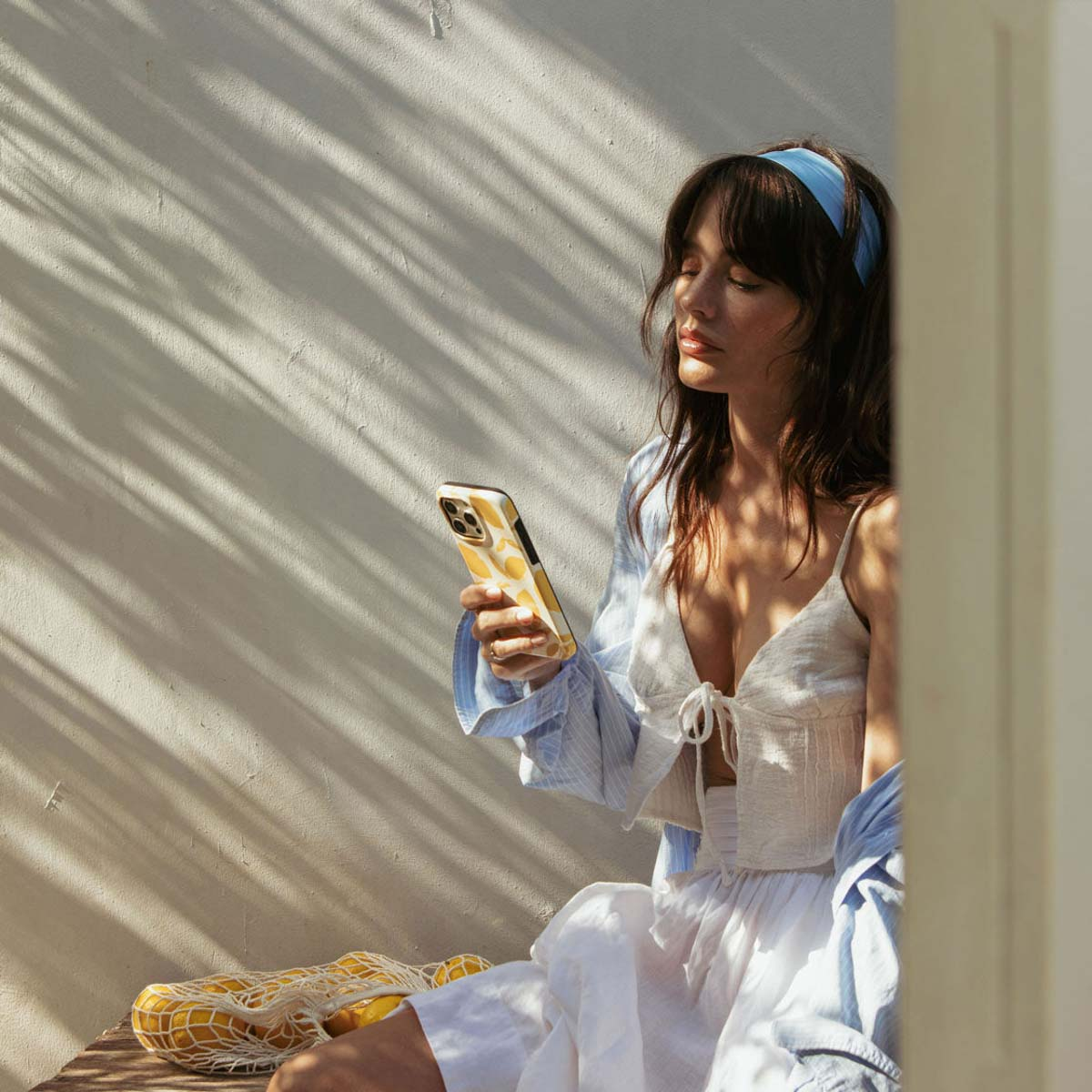 Une femme avec un bandeau bleu tient un téléphone à motifs de citrons. Elle porte un haut et une jupe blancs. Soleil et ombres sur le mur.