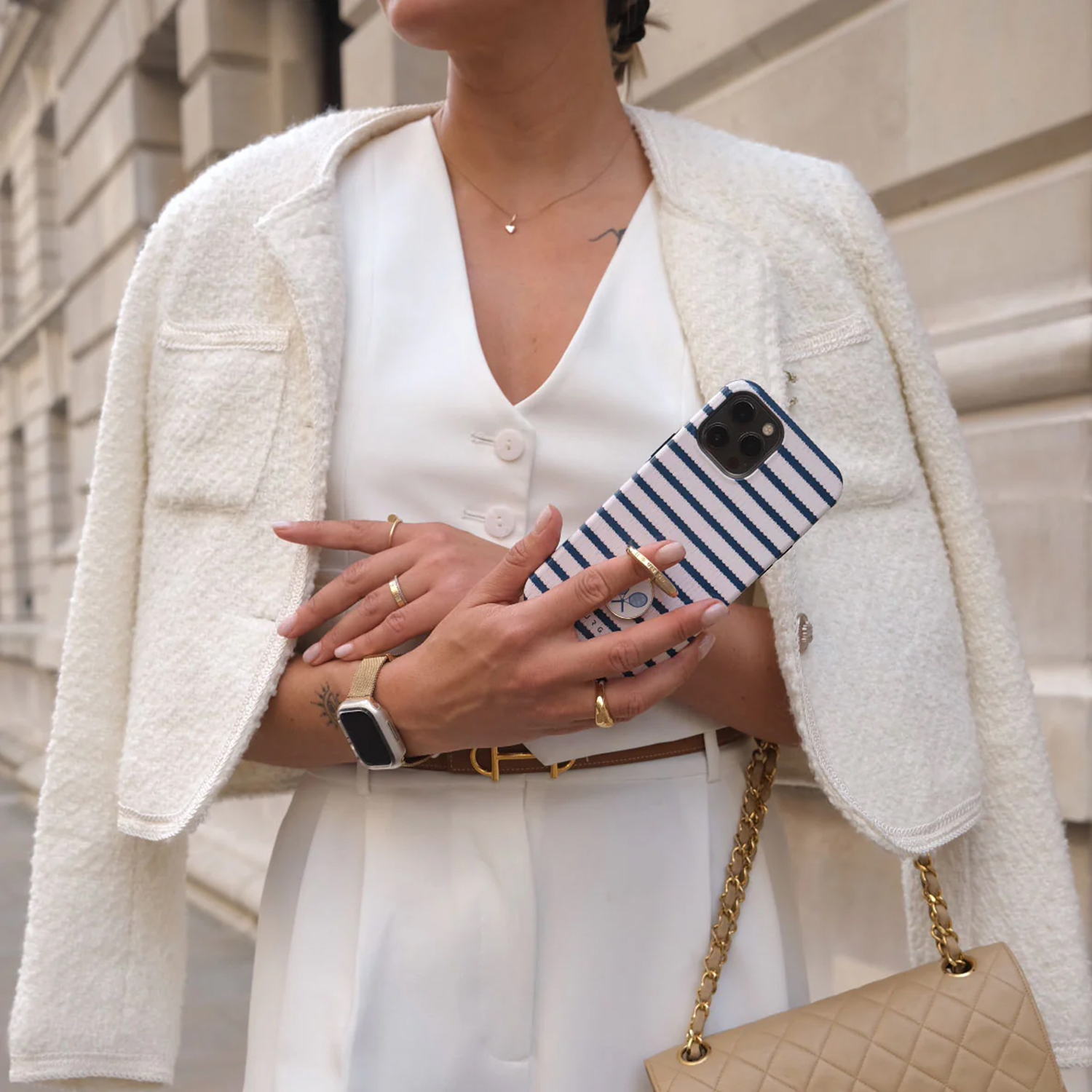 Vrouw in witte outfit houdt telefoon met gestreept hoesje vast. Ze draagt een jasje, ringen en een horloge. Een beige tas is zichtbaar.