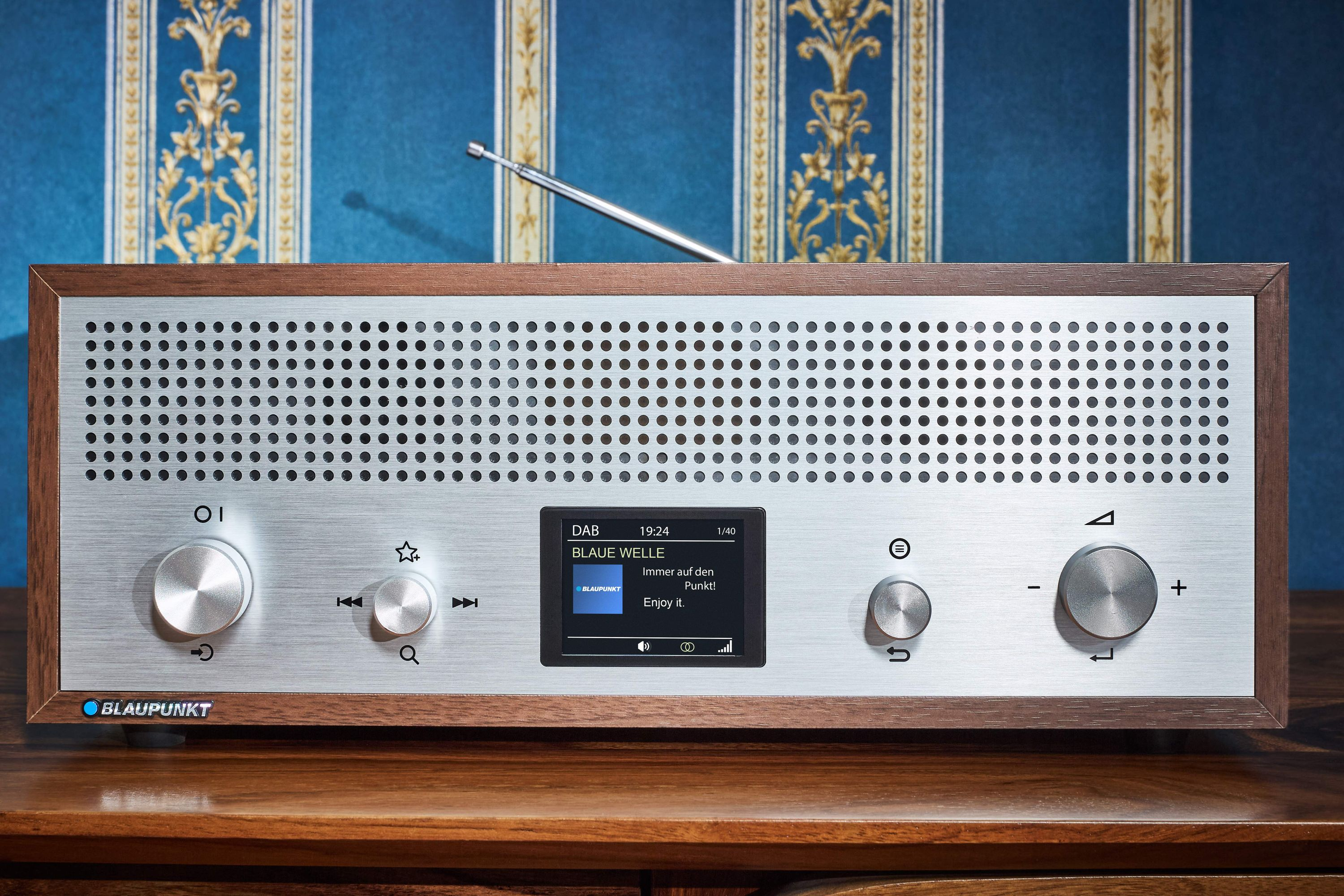 Ein Radio mit silberner Front, Holzrahmen und kleinem Display mit 'BLAUE WELLE'. Antenne ist ausgezogen.