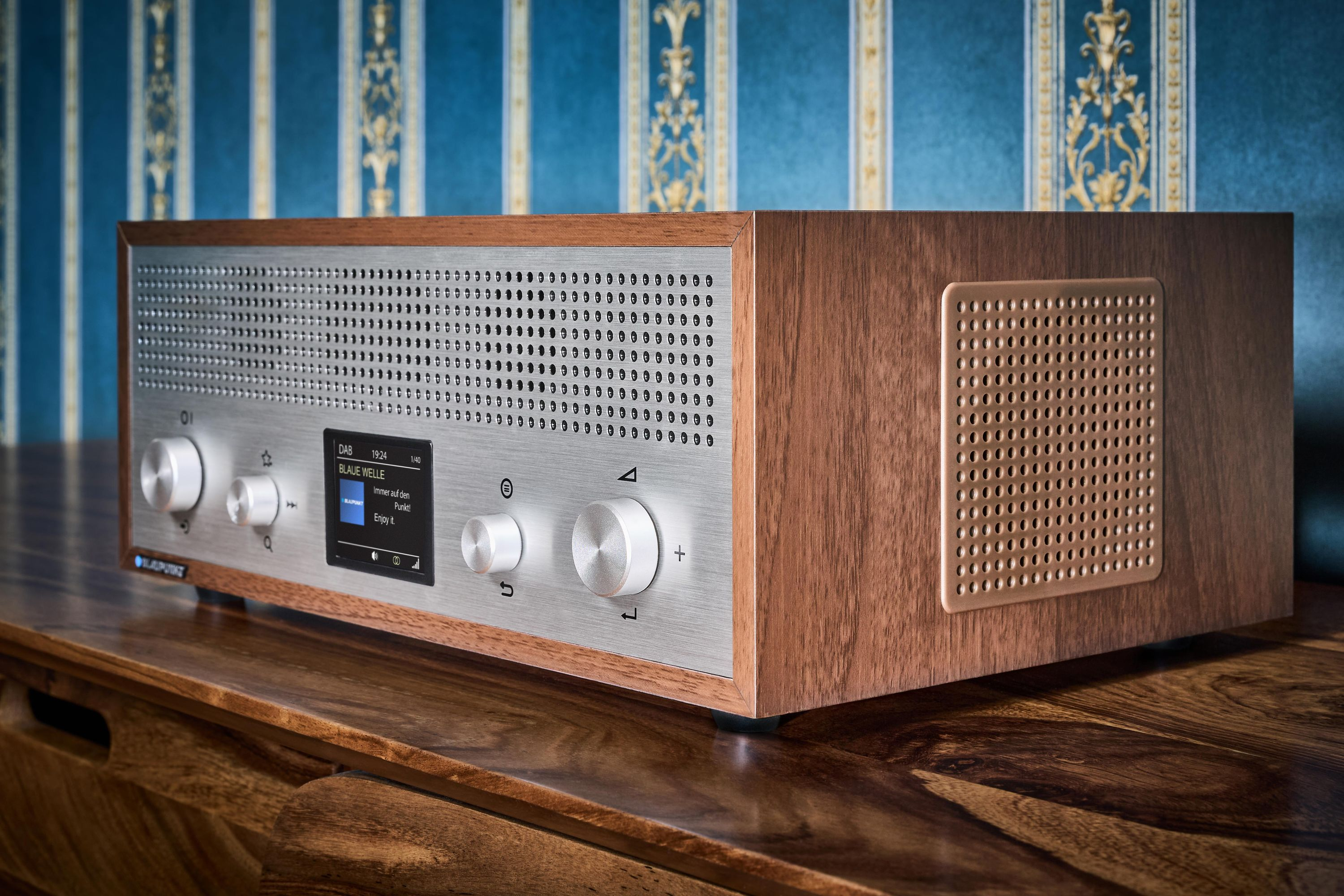 Ein Holzradio mit silberner Front und Bildschirm. Es steht auf einem Holztisch, im Hintergrund eine blau gemusterte Wand.