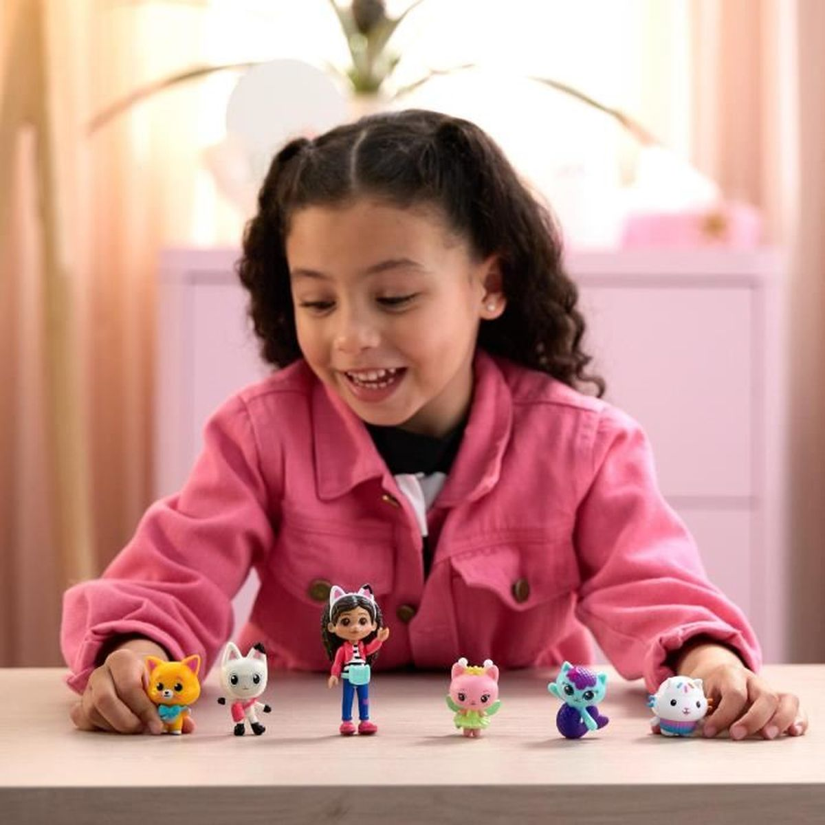 Ein Mädchen lächelt, umgeben von Gabby's Puppenhaus-Figuren. Die Spielzeuge zeigen katzenartige Charaktere. Der Hintergrund zeigt einen rosa Schrank.