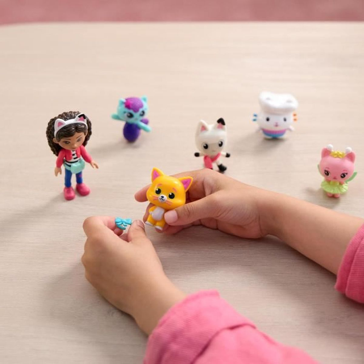 Kinderhände halten ein orangefarbenes Katzenspielzeug und eine blaue Schleife. Weitere Katzenspielzeuge und eine Puppe liegen auf dem Tisch.
