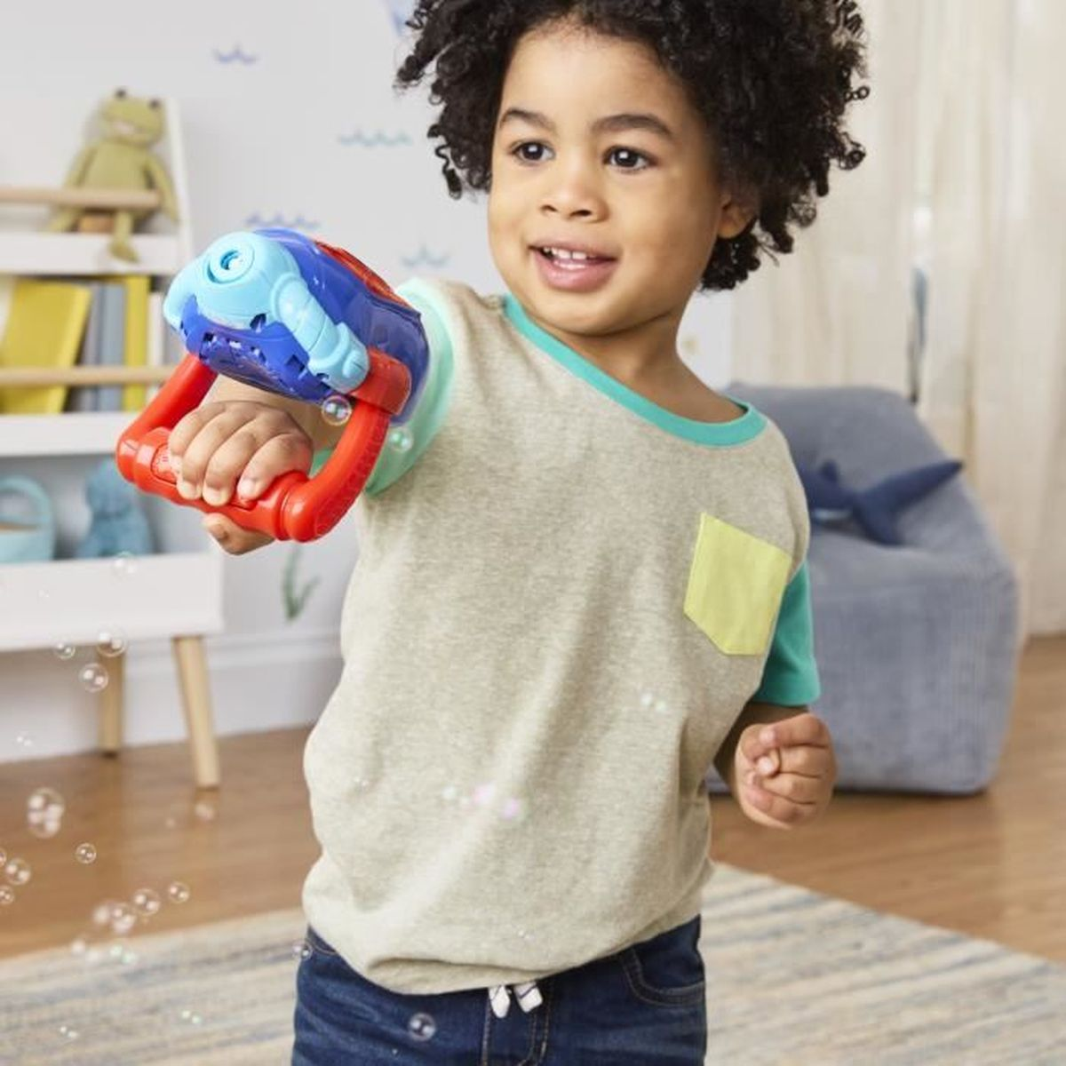 Un niño sonríe sosteniendo una pistola de juguete roja y azul, rodeado de burbujas. Lleva una camisa gris con un bolsillo amarillo.