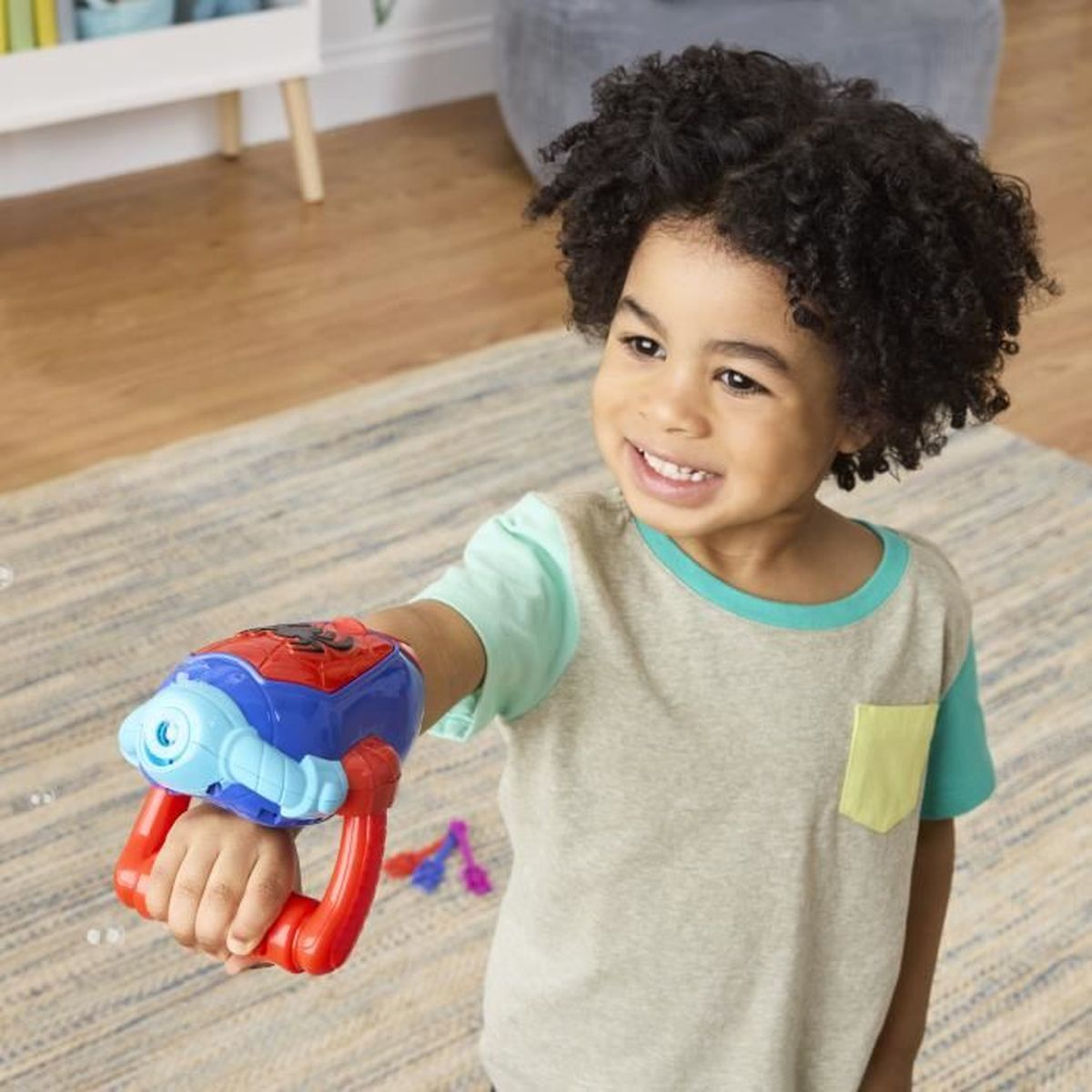 Un niño sonriente sostiene una pistola de juguete roja y azul. El niño lleva una camisa verde y beige. Una alfombra y suelo de madera están en el fondo.