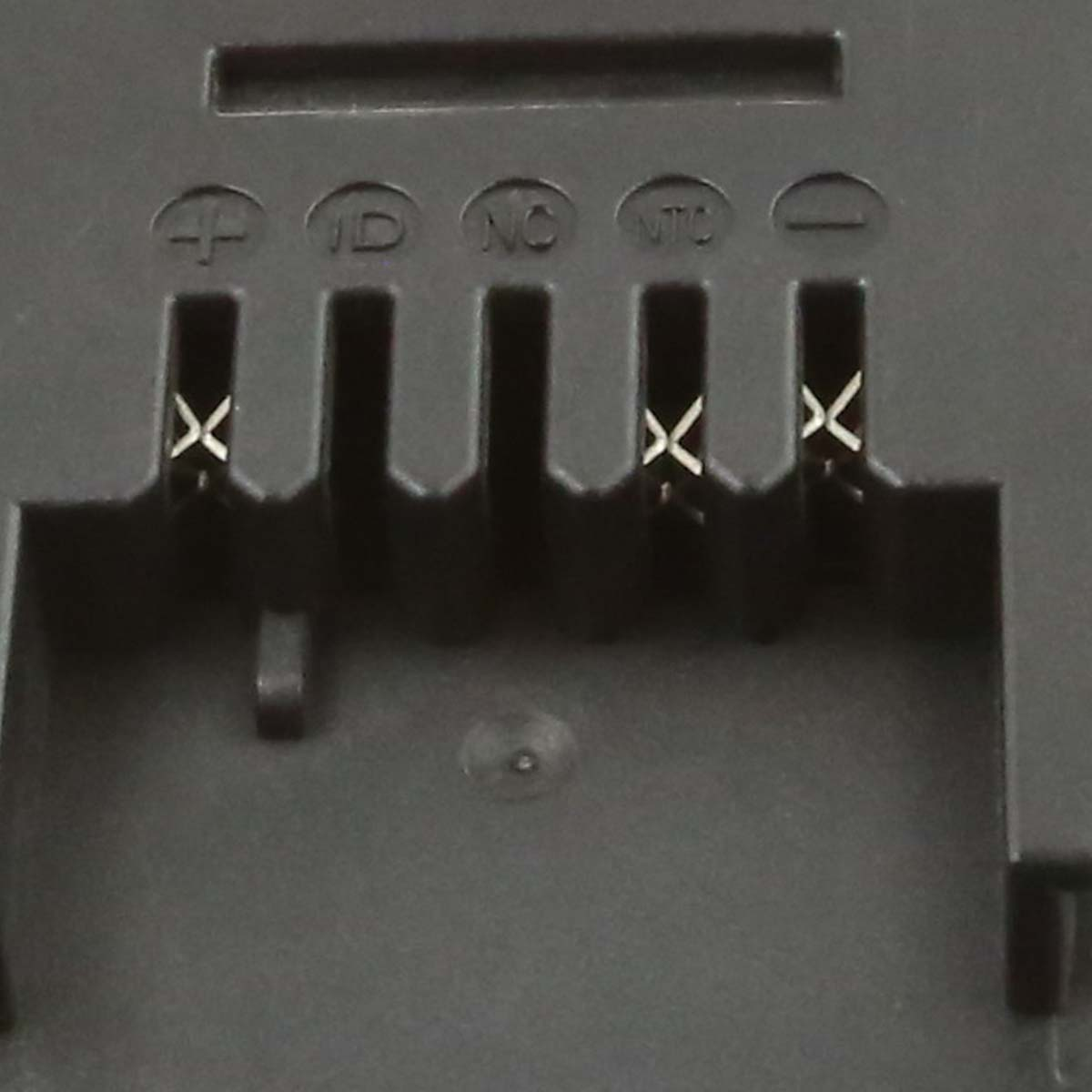 Nahaufnahme eines schwarzen Batteriefachs mit beschrifteten Anschlüssen: +, ID, NC, NTC und -. Metallkontaktpunkte sind sichtbar.