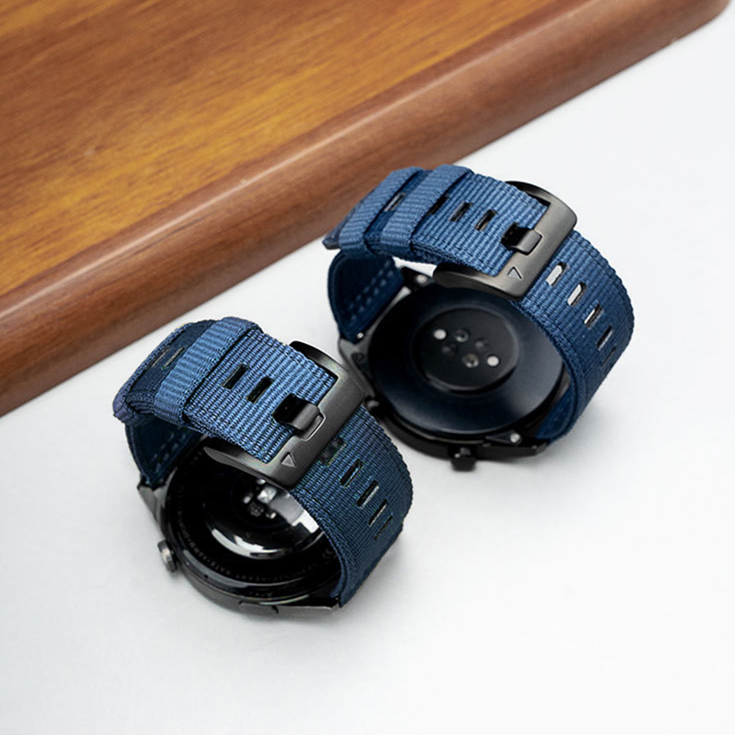 Due smartwatch neri con cinturini in tessuto blu. Uno è davanti all'altro. Sono su una superficie bianca con una tavola di legno.