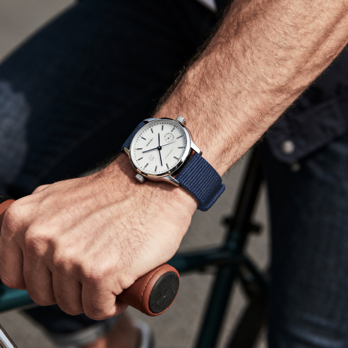 Arm einer Person mit Armbanduhr am Handgelenk. Die Uhr hat ein weißes Zifferblatt und ein blaues Armband. Die Person hält einen Fahrradlenker.