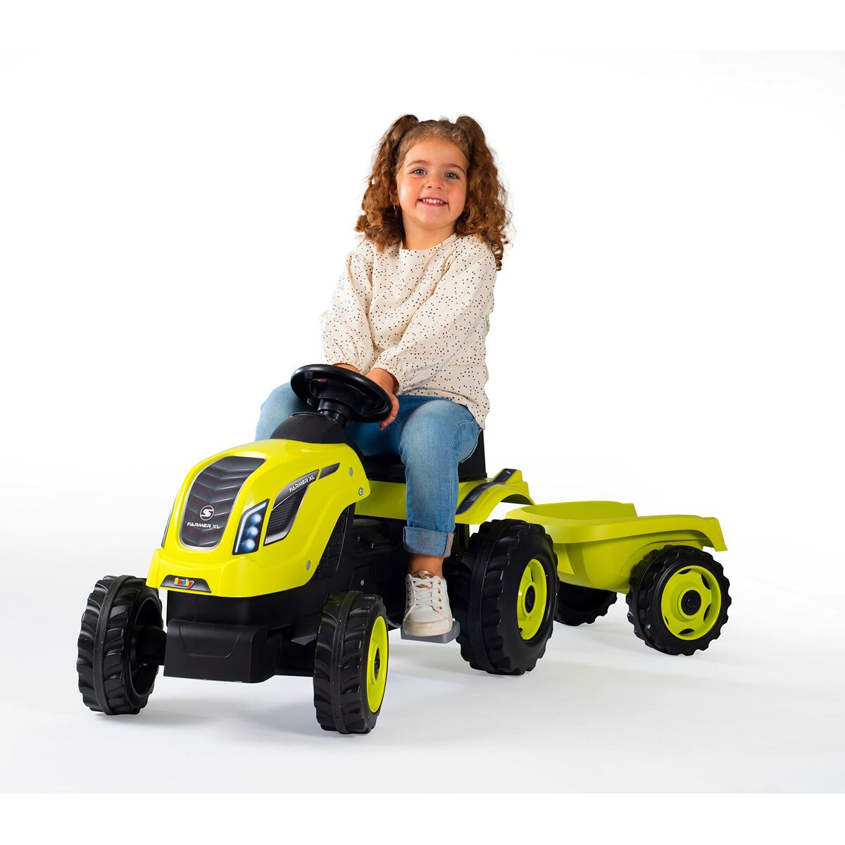 Ein junges Mädchen sitzt auf einem gelben Spielzeugtraktor mit Anhänger. Sie lächelt in die Kamera. Der Hintergrund ist weiß.