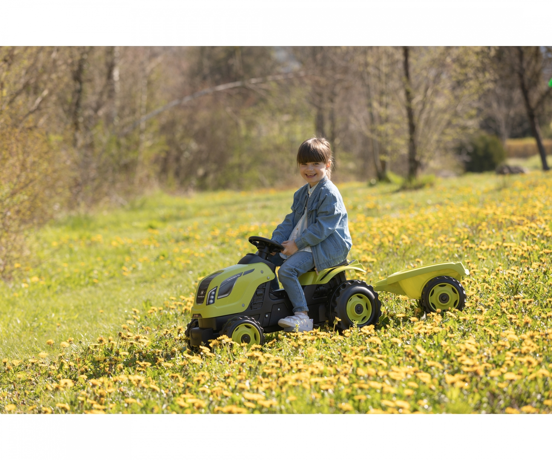 Ein Mädchen sitzt lächelnd auf einem grünen Spielzeugtraktor mit Anhänger in einem Feld mit gelben Blumen.