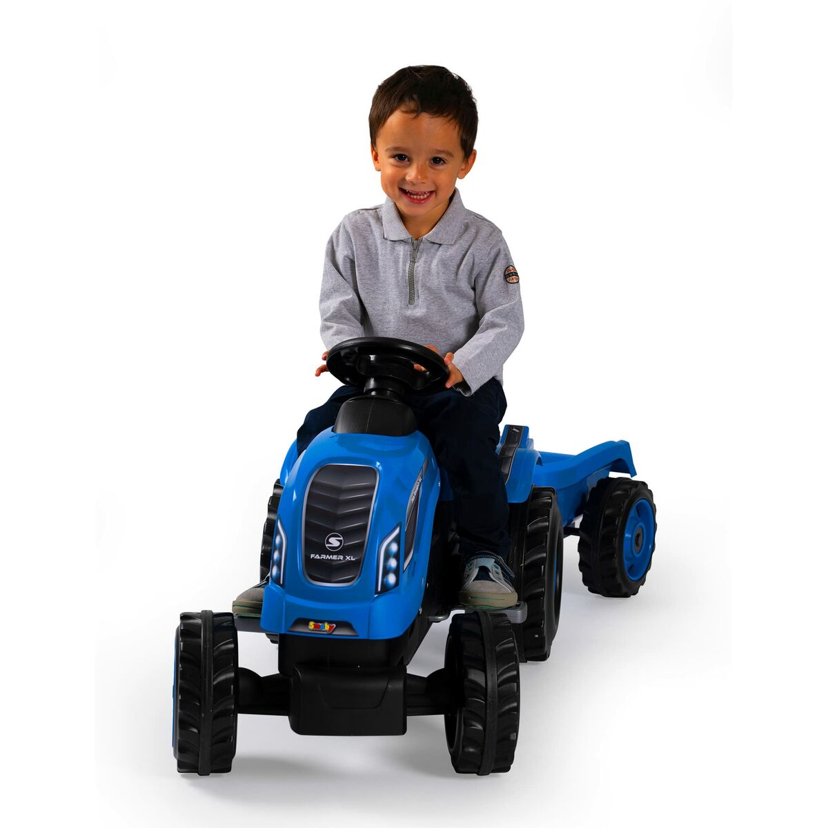 Ein Junge sitzt lächelnd auf einem blauen Spielzeugtraktor. Der Traktor hat große schwarze Reifen und steht auf weißem Hintergrund.