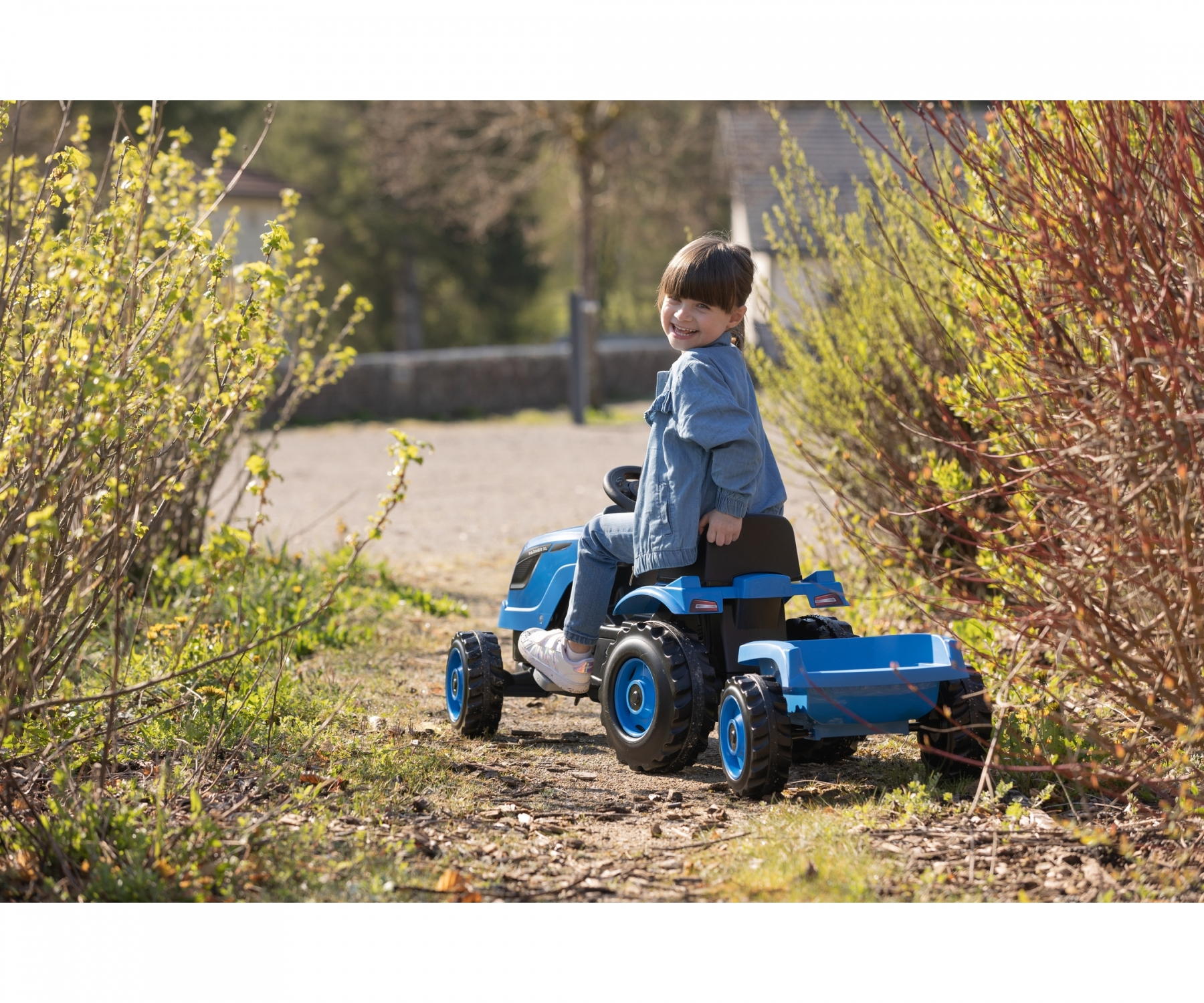 Ein Kind sitzt lächelnd auf einem blauen Spielzeugtraktor. Die Szene ist im Freien mit Büschen und einem verschwommenen Hintergrund.