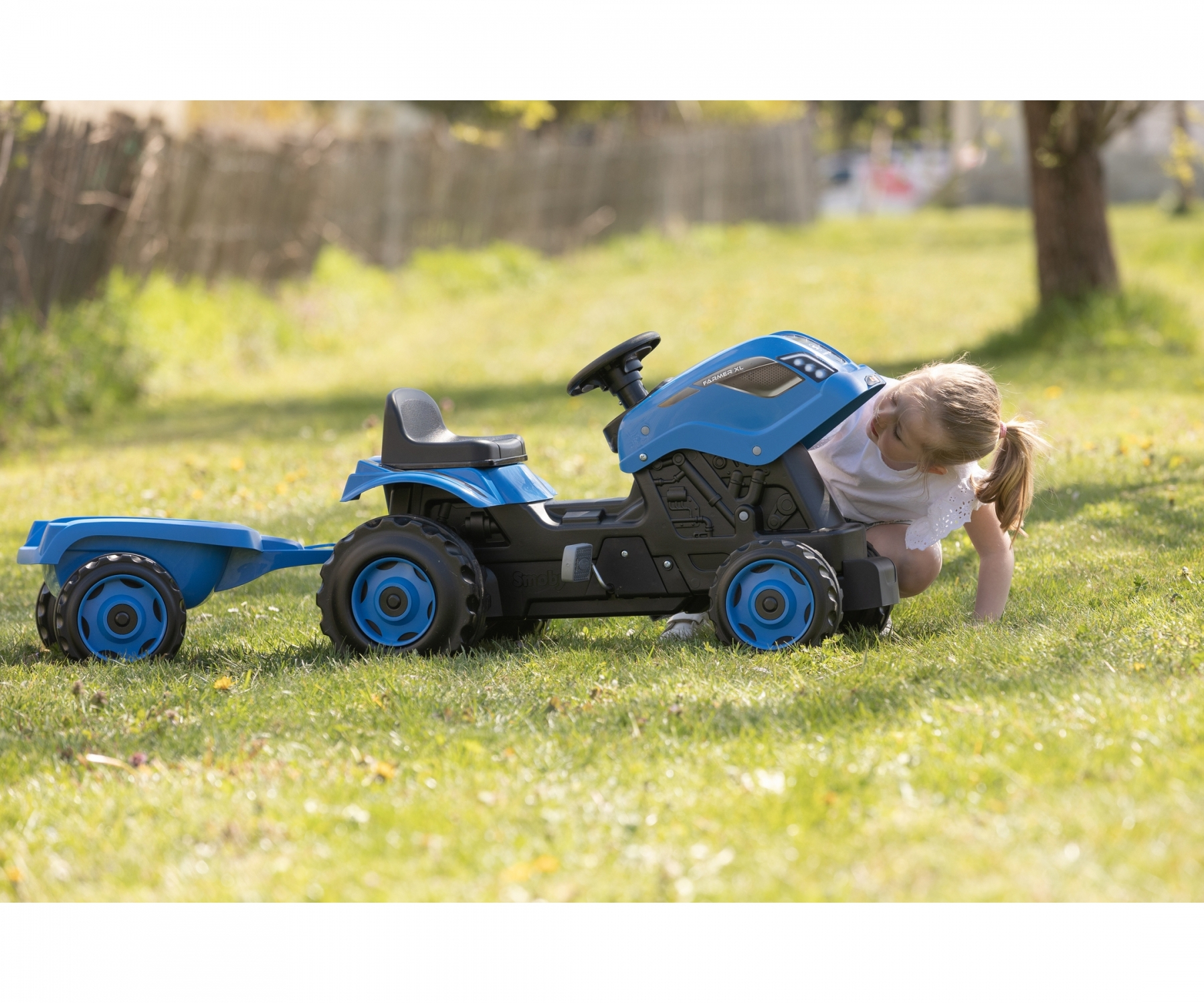 Ein Mädchen betrachtet einen blauen Spielzeugtraktor mit Anhänger auf Gras. Ein Holzzaun und Bäume sind im Hintergrund.
