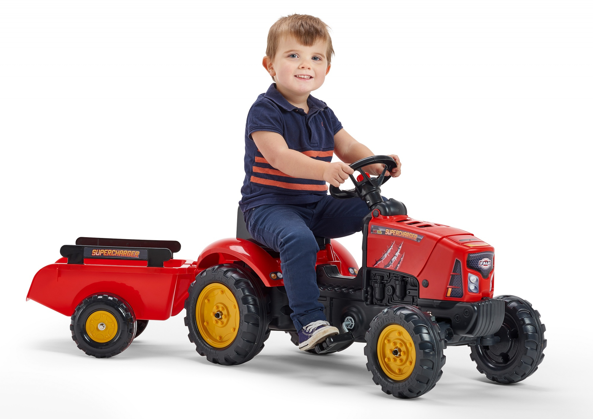 Ein lächelnder Junge sitzt auf einem roten Spielzeugtraktor mit Anhänger. Er trägt ein blaues Hemd und Jeans. Der Traktor hat gelbe Räder.