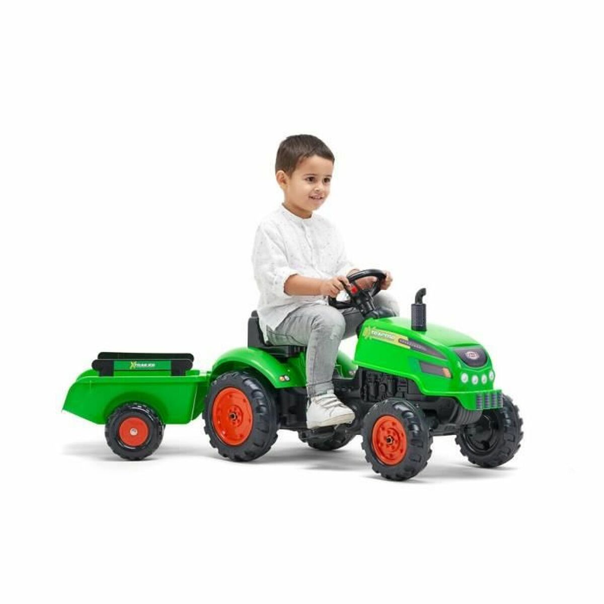 Ein Kind sitzt auf einem grünen Spielzeugtraktor mit Anhänger. Der Traktor hat orangefarbene Räder. Das Kind lächelt und trägt ein weißes Hemd.