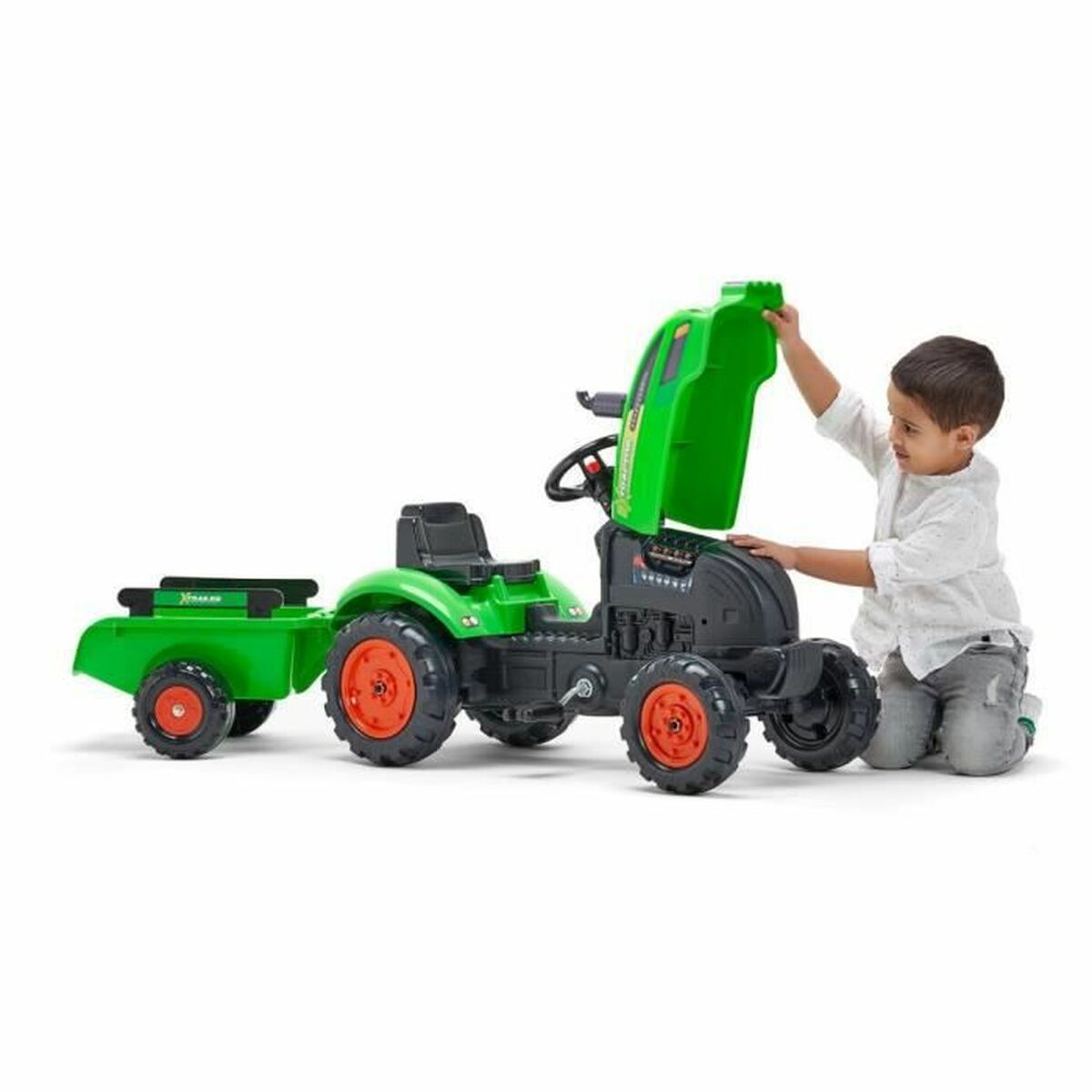 Ein kleiner Junge kniet neben einem grünen Spielzeugtraktor mit Anhänger und hebt die Motorhaube an. Der Traktor hat orangefarbene Räder.