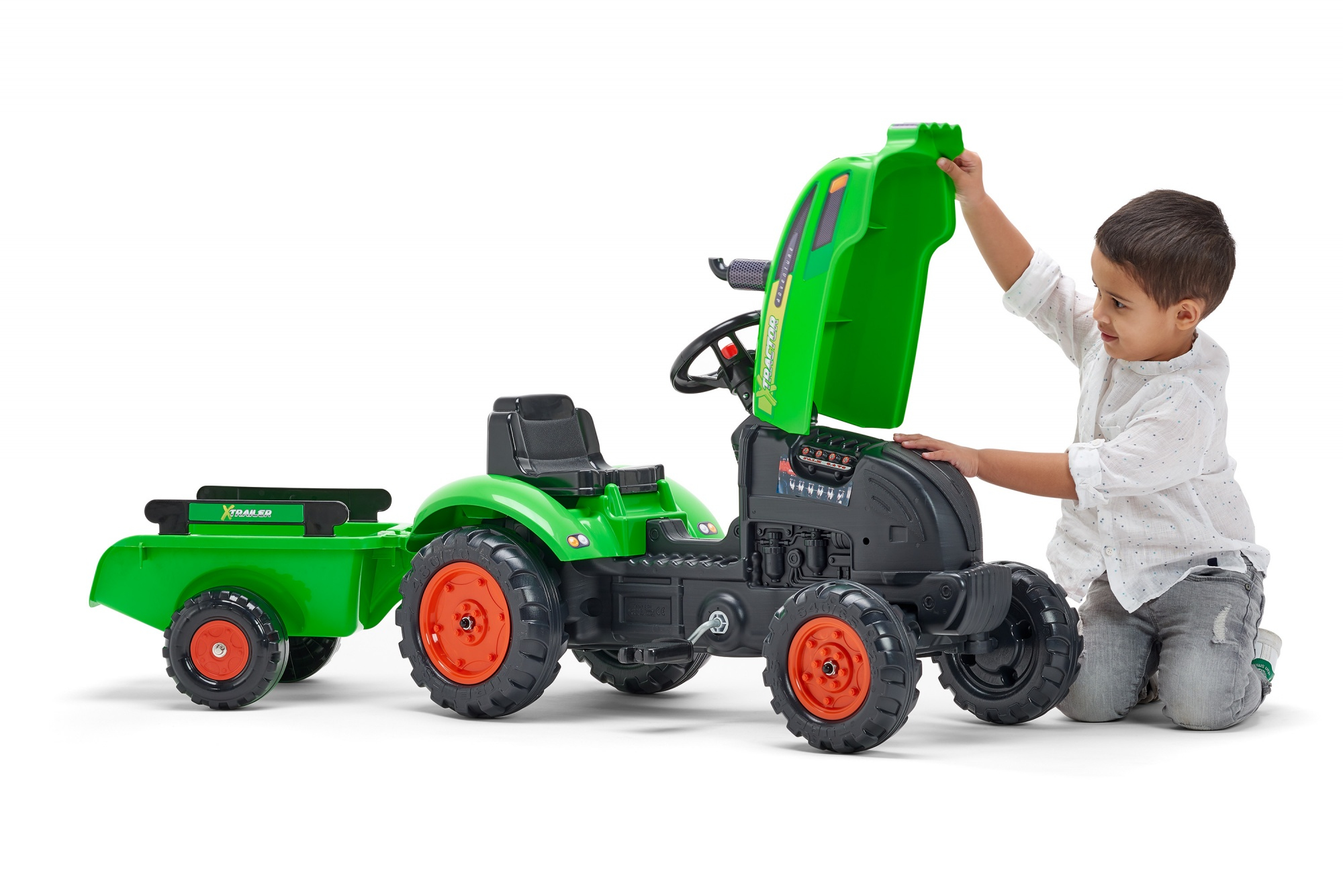 Ein kleiner Junge kniet neben einem grünen Spielzeugtraktor mit Anhänger. Er hebt die Motorhaube an. Der Traktor hat orangefarbene Räder.