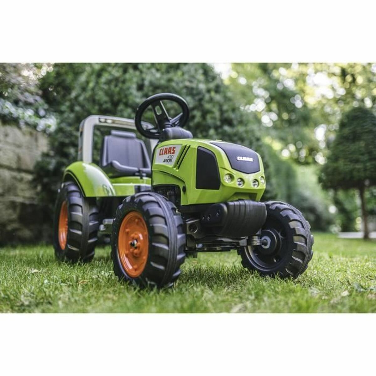 Ein grüner Spielzeugtraktor auf Gras, mit orangefarbenen Rädern und einem schwarzen Lenkrad, vor dem Hintergrund grüner Büsche und Bäume.
