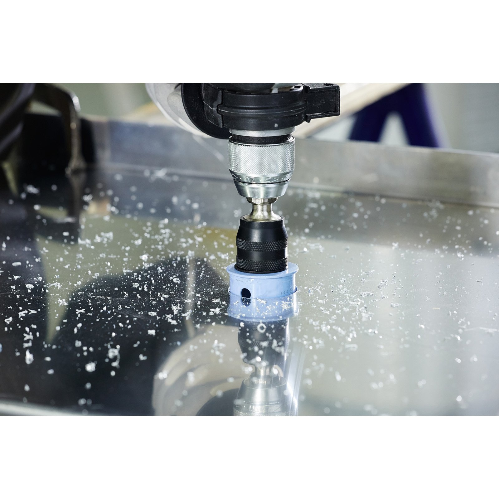 Eine Ständerbohrmaschine bohrt ein Loch in ein Metallblech. Metallspäne liegen um den Bohrer herum verstreut. Eine blaue Führung ist angebracht.