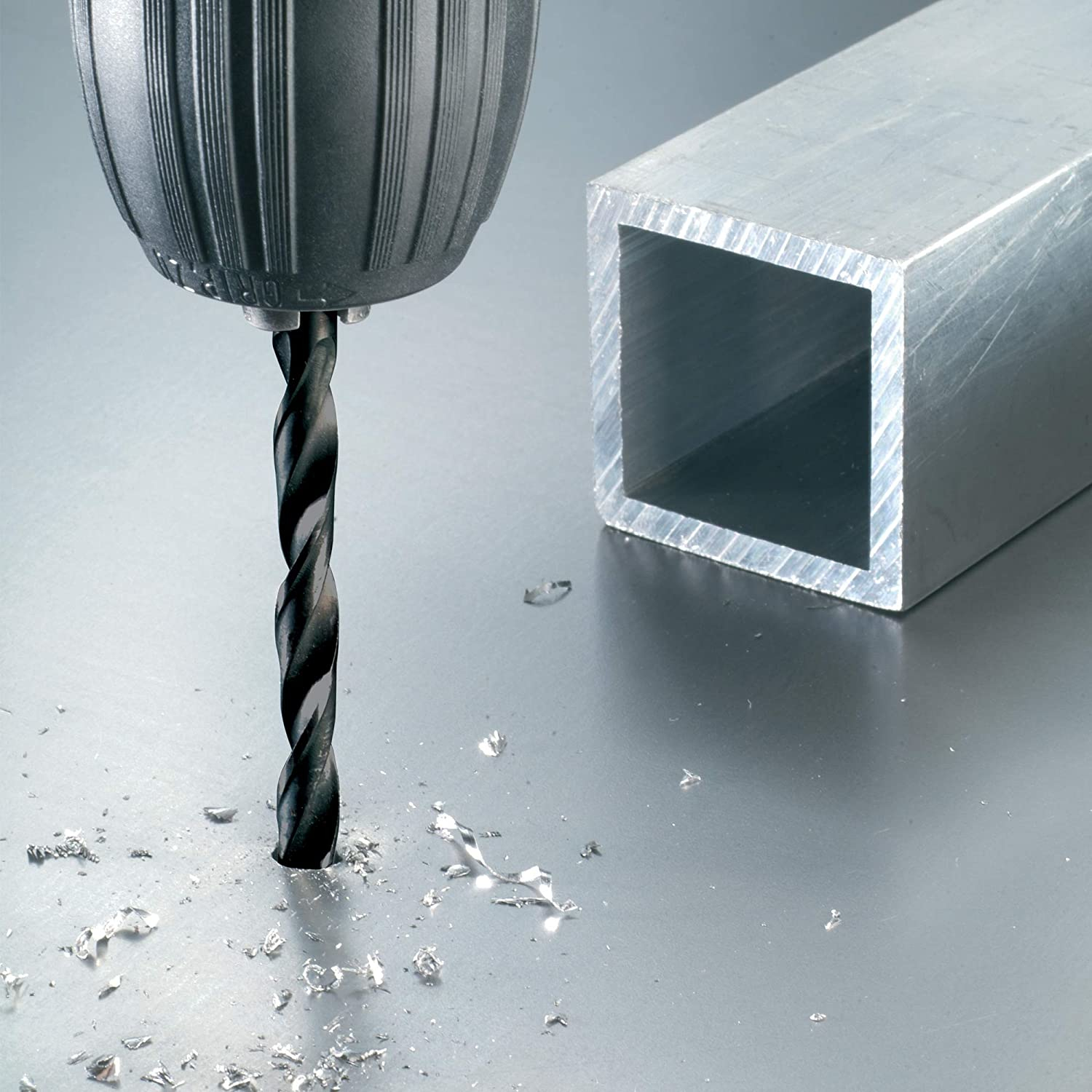 Ein Bohrer bohrt in eine Metalloberfläche. Ein quadratisches Metallrohr befindet sich im Hintergrund. Metallspäne liegen um den Bohrer herum.