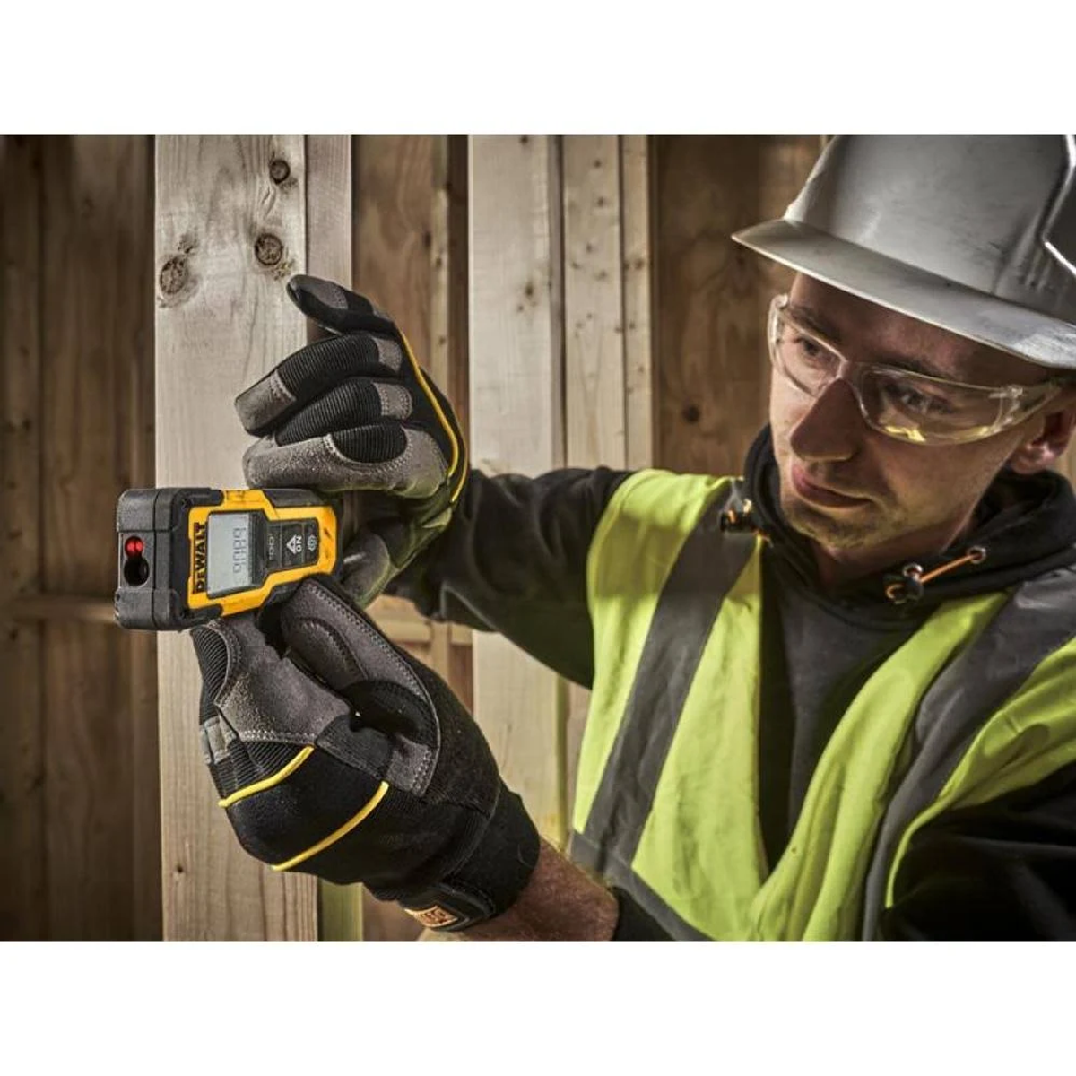 Ein Arbeiter mit Handschuhen, Brille und Helm benutzt ein Laser-Entfernungsmessgerät an Holzbalken.