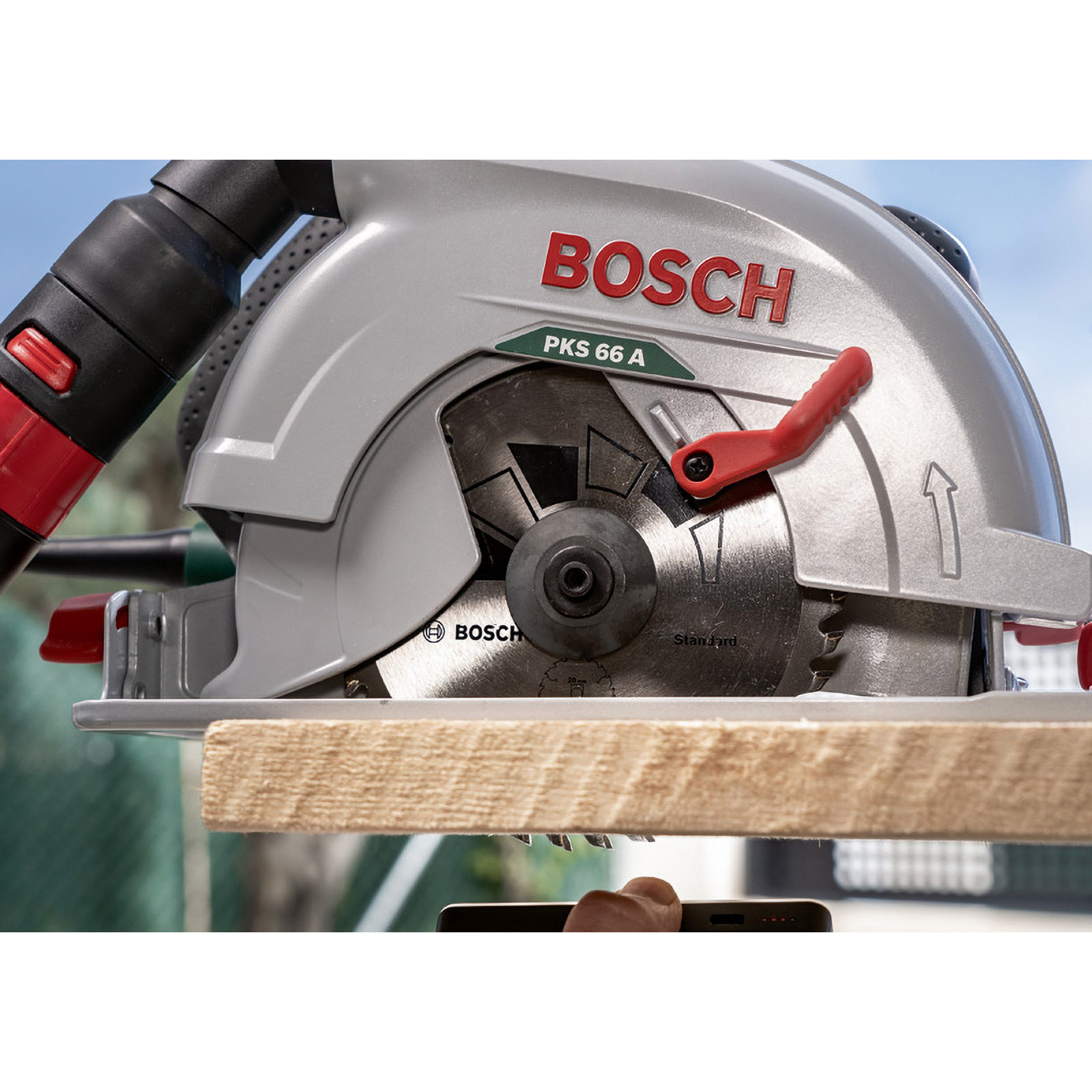 Eine Bosch PKS 66 A Kreissäge schneidet Holz. Rote und graue Details. Himmel im Hintergrund.