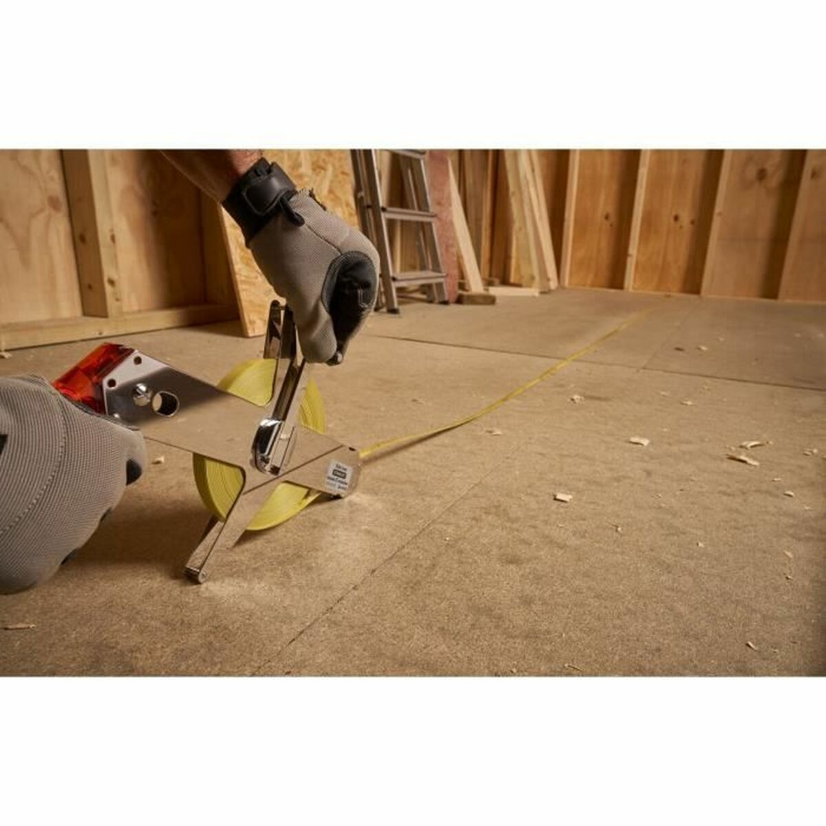 Eine Person verwendet ein Metall-Messband auf einem Boden. Ein gelbes Band erstreckt sich über den Boden. Holzständer und eine Leiter sind im Hintergrund.