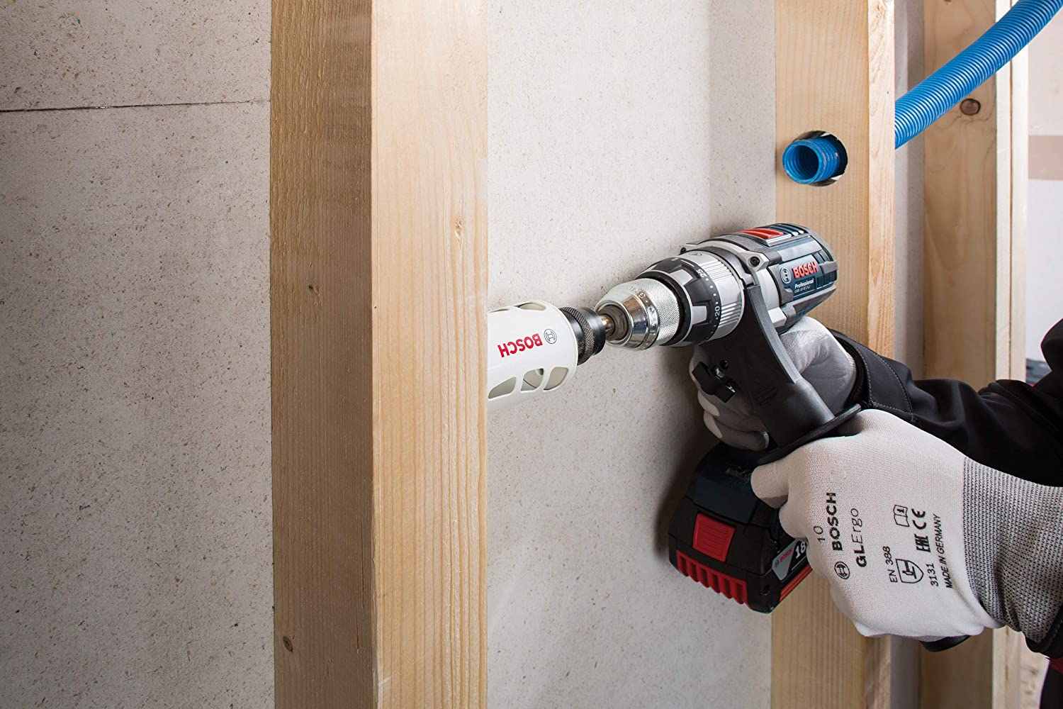 Eine Person benutzt einen Bosch-Bohrer mit einer Lochsäge, um in eine Holzwand zu bohren. Im Hintergrund ist ein blaues Rohr zu sehen.