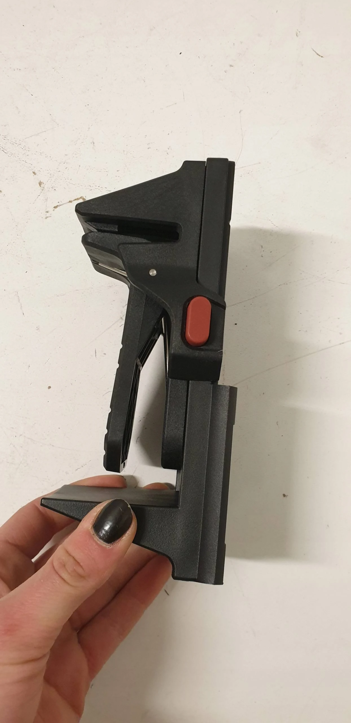 Ein schwarzes Werkzeug mit rotem Knopf wird von einer Hand mit schwarzem Nagellack gehalten. Es liegt auf einer weißen Oberfläche.