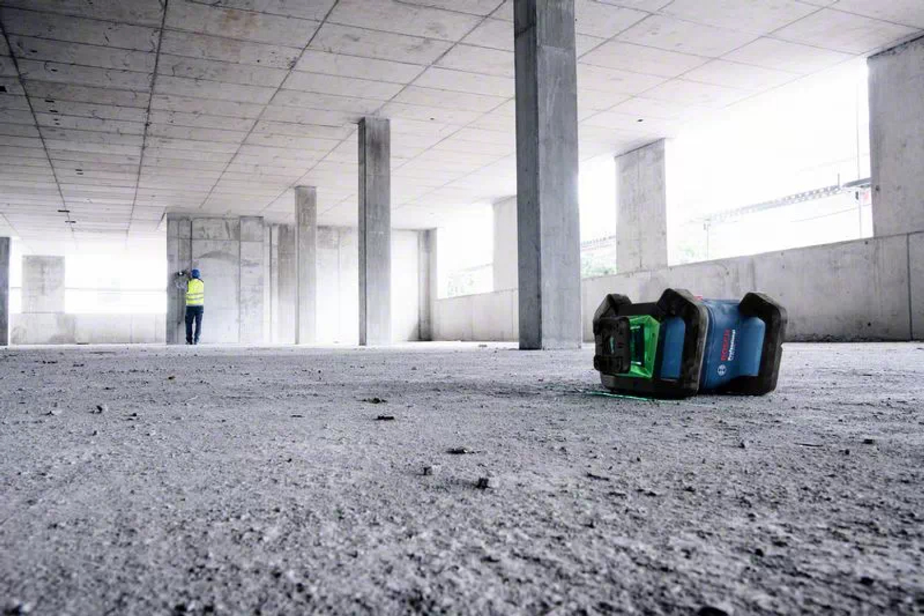 Eine Baustelle zeigt einen Arbeiter und ein Laser-Niveau. Betonpfeiler tragen eine Decke. Der Boden ist rau.