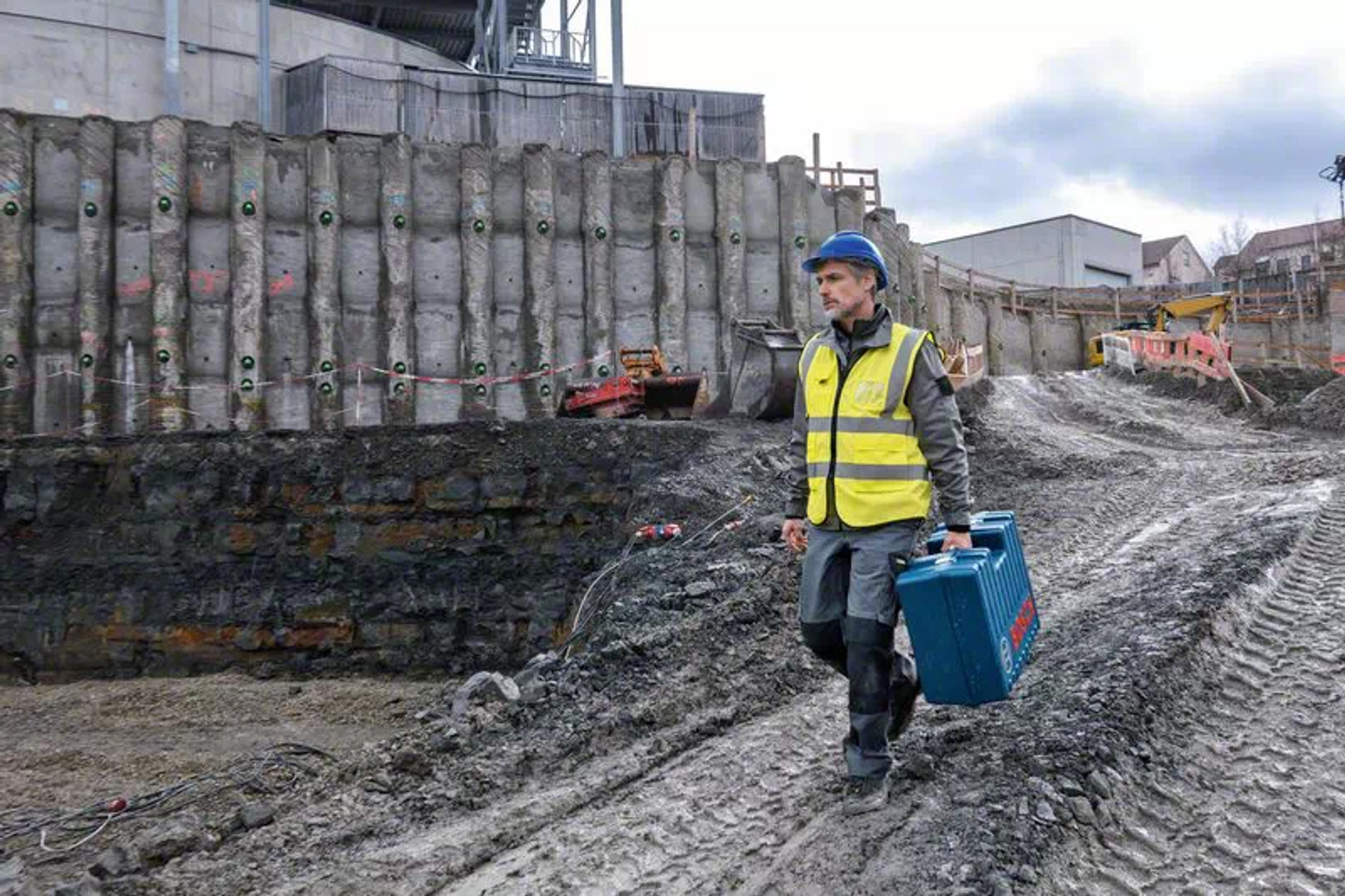 Ein Bauarbeiter in gelber Weste und blauem Helm trägt einen blauen Koffer auf einer schlammigen Baustelle mit einer Stützmauer.