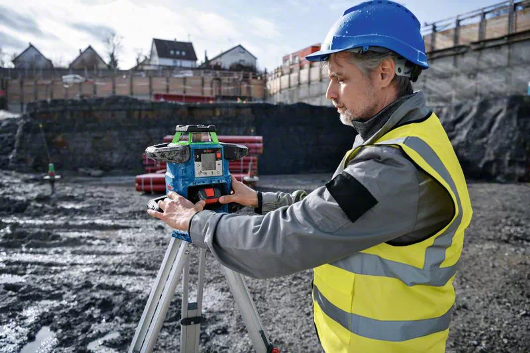 Ein Bauarbeiter mit blauem Helm und gelber Weste benutzt einen Laser-Nivellierer auf einem Stativ. Häuser und Baustelle im Hintergrund.